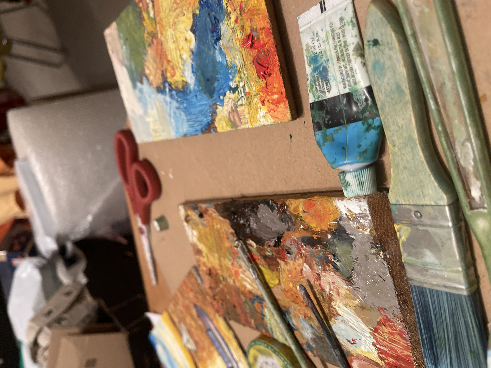 The image shows an artist's workspace. There are two small painted canvases, one with a landscape featuring green, yellow, blue, and red tones, and the other with an abstract scene of black, grey, orange, and white. A squeezed tube of blue and green paint lies near the top right corner of the image. Two paintbrushes, heavily used and stained with paint, are positioned vertically to the right of the paint tube. A pair of red scissors is placed diagonally across the frame. Background details are obscured, but bubble wrap and boxes are seen in the upper left. The location of Maidstone, United Kingdom is not evident in the image.