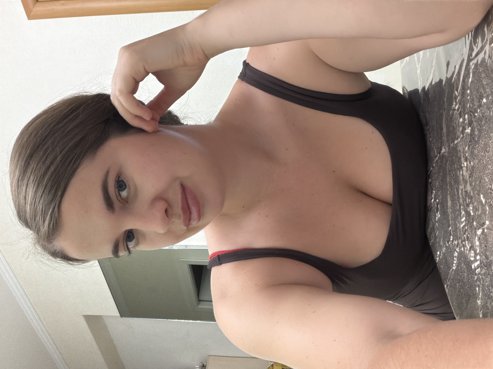 A woman is shown in a selfie. She is wearing a black tank top. Her hand is near her head. The background appears to be a room with neutral tones. The location is unknown and cannot be determined from the image.