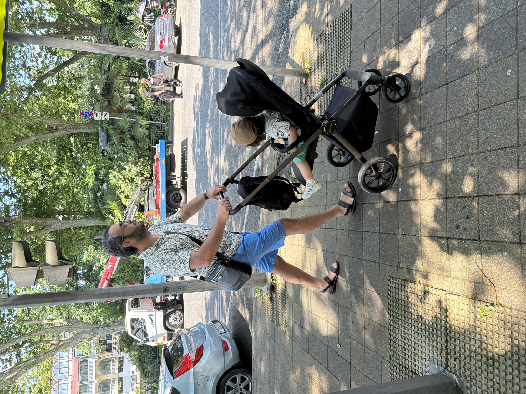 A man wearing a patterned shirt and blue shorts is pushing a stroller with a toddler in it. The man has a beard and is wearing glasses and sandals. The toddler, wearing a cap and a t-shirt with a cartoon character, is seated in the stroller. A black bag is attached to the stroller.
The scene appears to be a sunny day, as indicated by the shadows cast on the pavement. The location seems to be a city street or sidewalk, with trees and buildings visible in the background. There are also several vehicles, including a car, a truck, and a crane truck, suggesting an urban or semi-urban environment. The presence of a stroller and a young child implies a family outing. The overall mood appears to be relaxed and casual.
The image seems to be taken in Sochi, Russia, based on the context provided. There are no other visible text elements in the image.