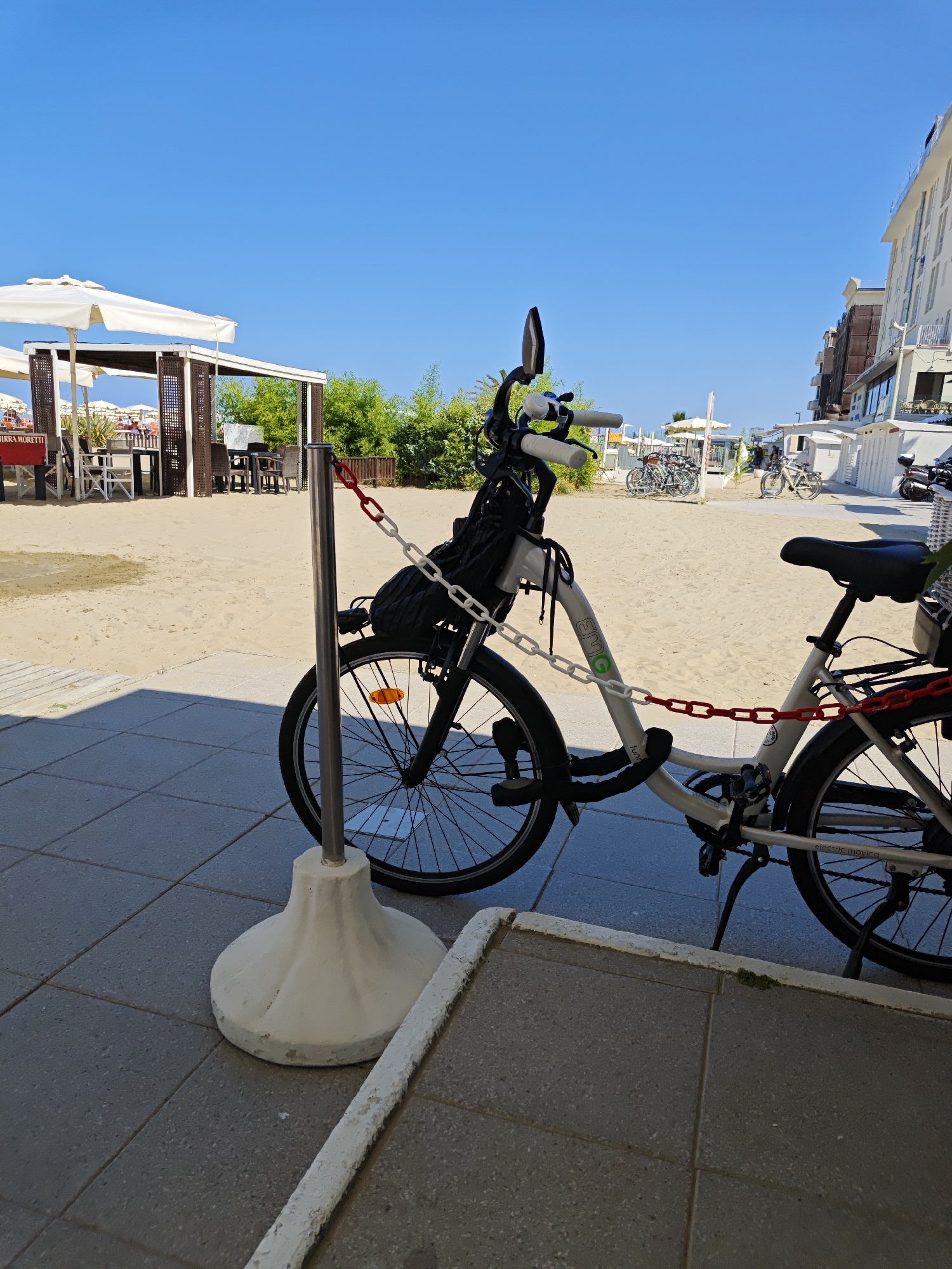 A silver electric bicycle, secured with a black lock and a red and white chain, is parked on a paved area next to a sandy beach. The bicycle has a black basket on the handlebars and a white basket on the rear rack. In the background, there are white umbrellas, tables, and chairs on the beach, suggesting a seaside cafe or restaurant. A multi-story building with white balconies is visible on the right side of the image. The sky is clear and blue. The location is the beach in Rimini, Italy.