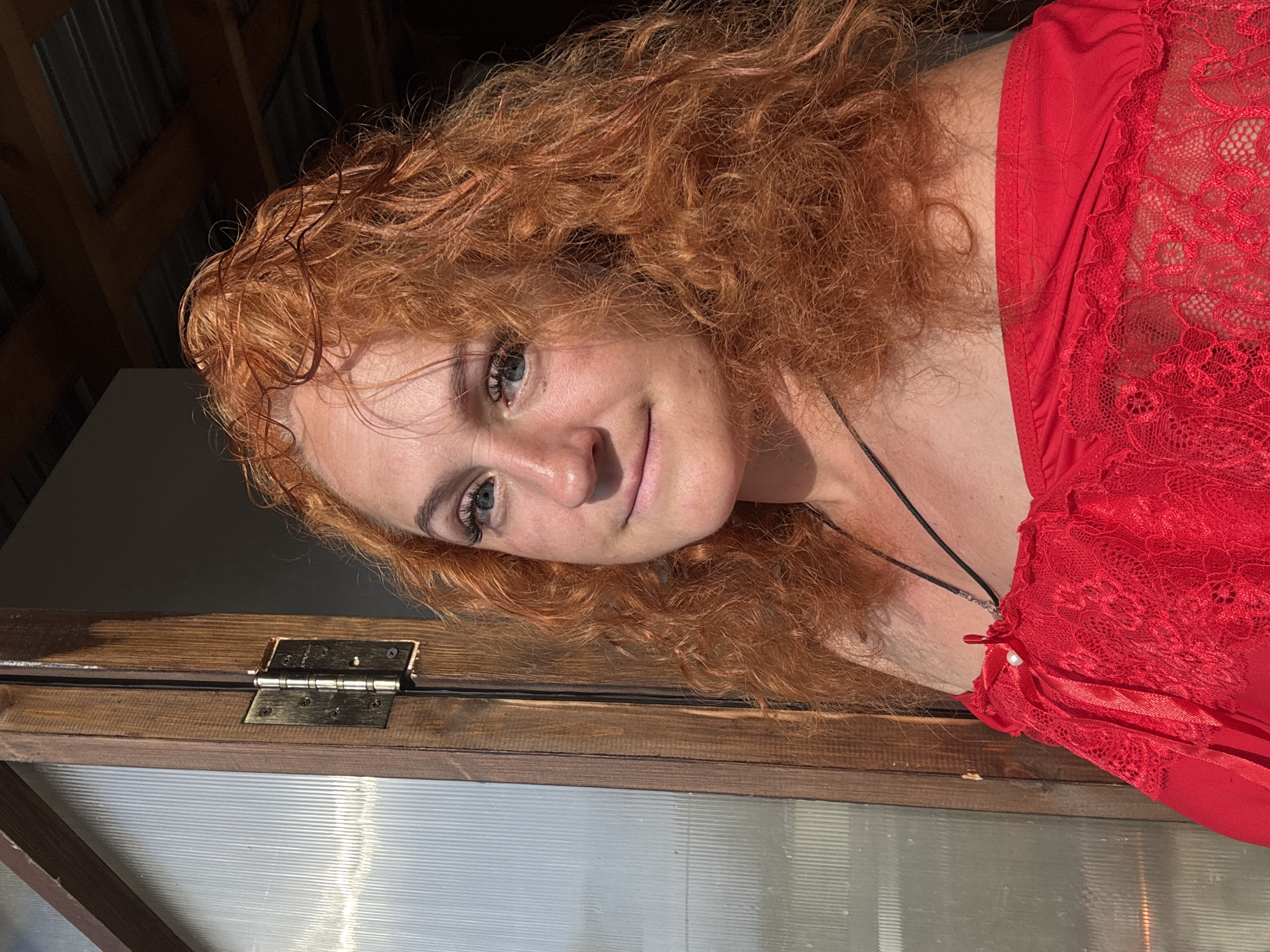 A woman with curly red hair and blue eyes is photographed outdoors, in the city of Chaikovsky, Russia. She is wearing a red off-the-shoulder top with lace trim. The woman is looking towards the camera, with a slight smile. Behind her, a wooden door with a glass panel is visible, and to the right, a metal roof structure can be seen. The lighting suggests it is daytime, with sunlight casting shadows.