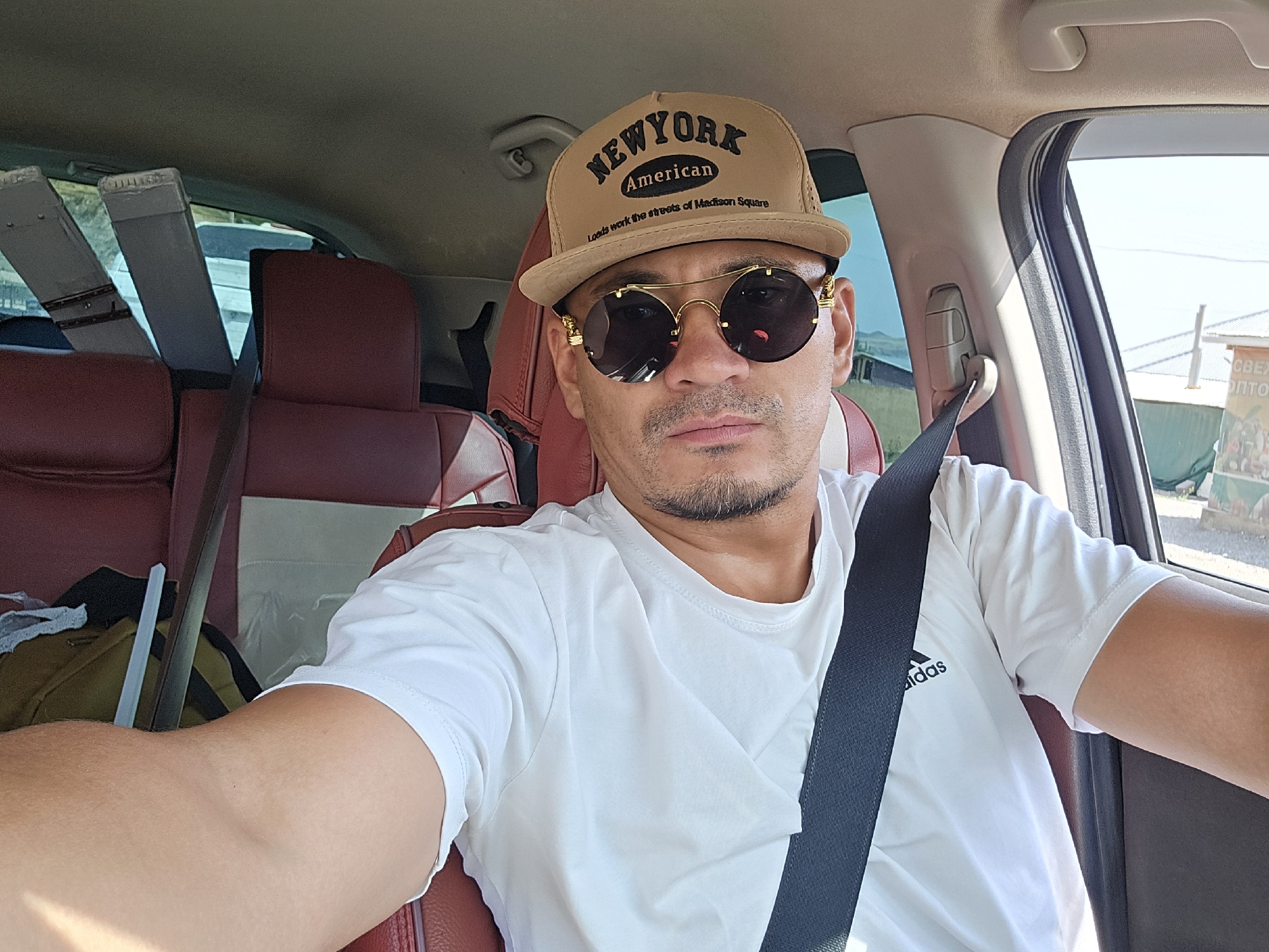 A man wearing a tan baseball cap with "NEW YORK AMERICAN" embroidered on it and gold-rimmed sunglasses is taking a selfie in the driver's seat of a car. He is wearing a white t-shirt with an Adidas logo on the left side. The car has red leather seats and a dark seatbelt is visible across his chest. In the background, through the car windows, a dry, dusty landscape is visible, with some buildings in the distance. The scene appears to be in an open area, possibly a rural or semi-urban setting. The presence of a sign with Cyrillic script in the background suggests the location is in a Russian-speaking region, consistent with Kızıl-Tokoy, Kyrgyzstan.