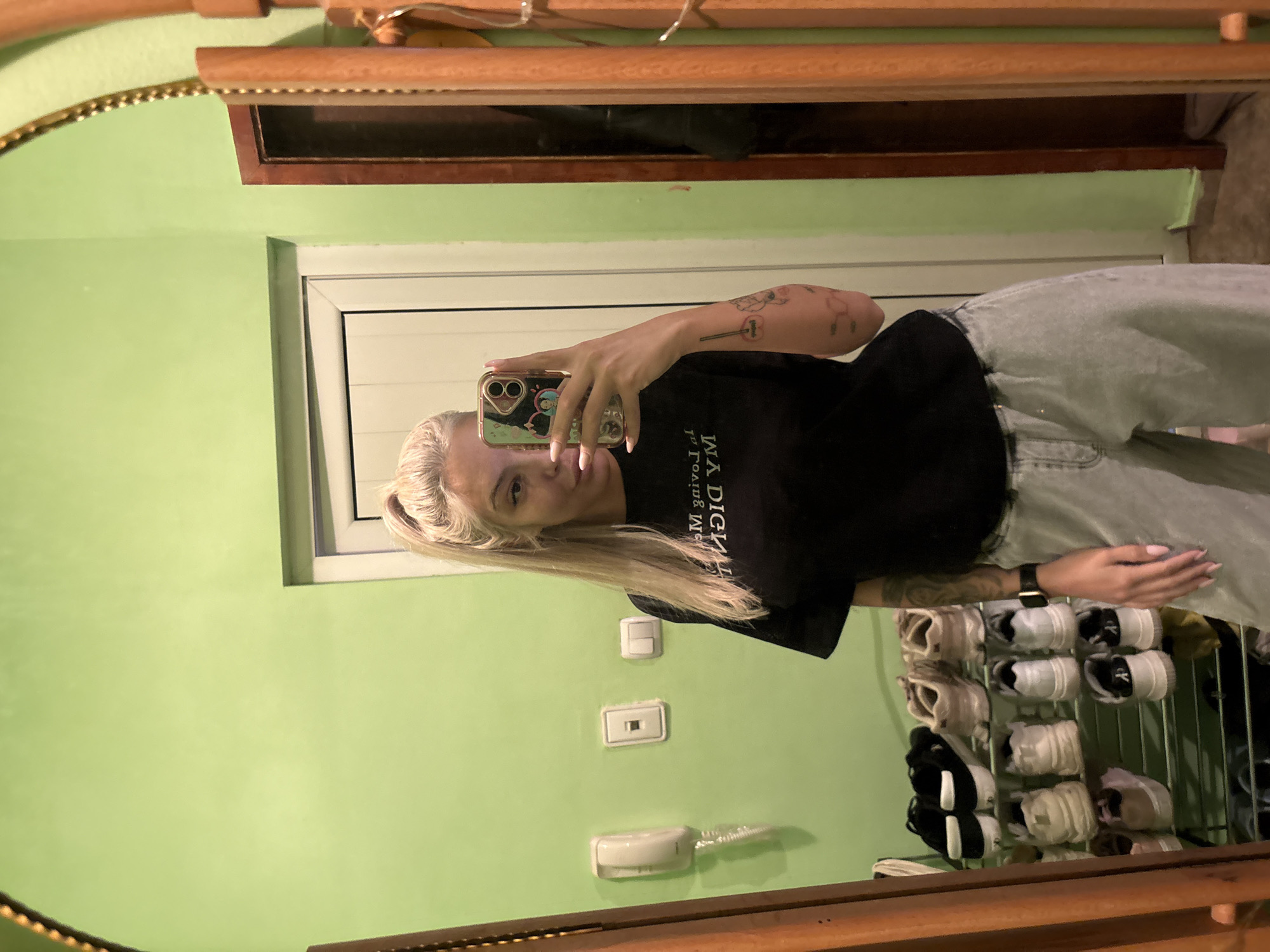 A blonde woman is taking a selfie in a mirror. She is wearing a black t-shirt and light-colored jeans. Her right arm is extended, holding a phone with a patterned case. She has tattoos on both arms. Behind her, there is a light green wall with electrical outlets and a white telephone. A shoe rack filled with sneakers is also visible. The mirror has a decorative wooden frame. The location is likely an apartment in Plovdiv, Bulgaria, given the context of the user's request.