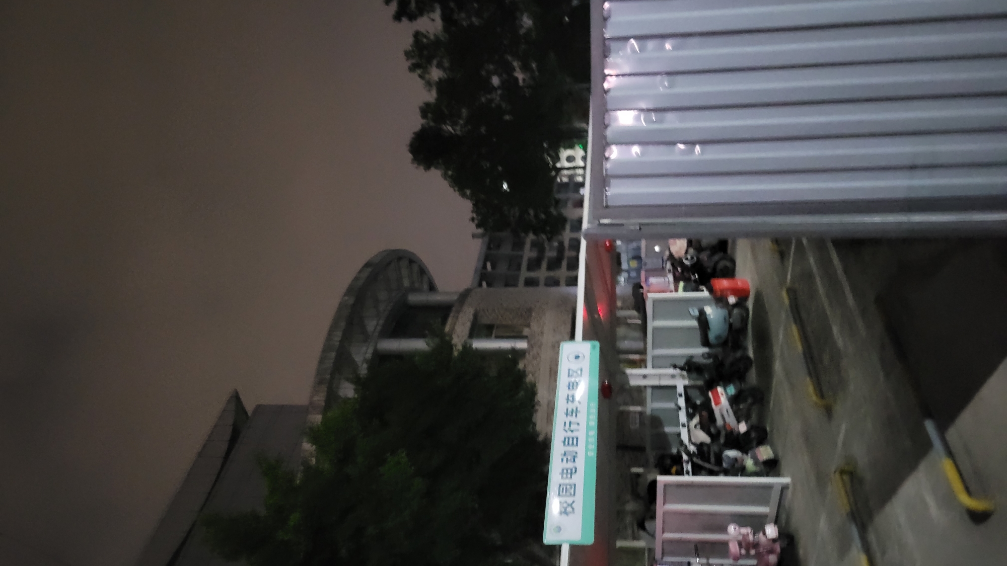 Captured from an elevated vantage point in Yuzhong District, China, the image presents a night scene focusing on a bicycle parking area. A green and white sign with Chinese characters identifies the location, adjacent to which are parked bicycles and electric scooters. The scene appears to be partially enclosed, with a roller shutter door visible at the top of the frame. Trees and architectural structures, including a building with a distinctive arched design, are visible in the background. The sky is overcast, indicating nighttime.