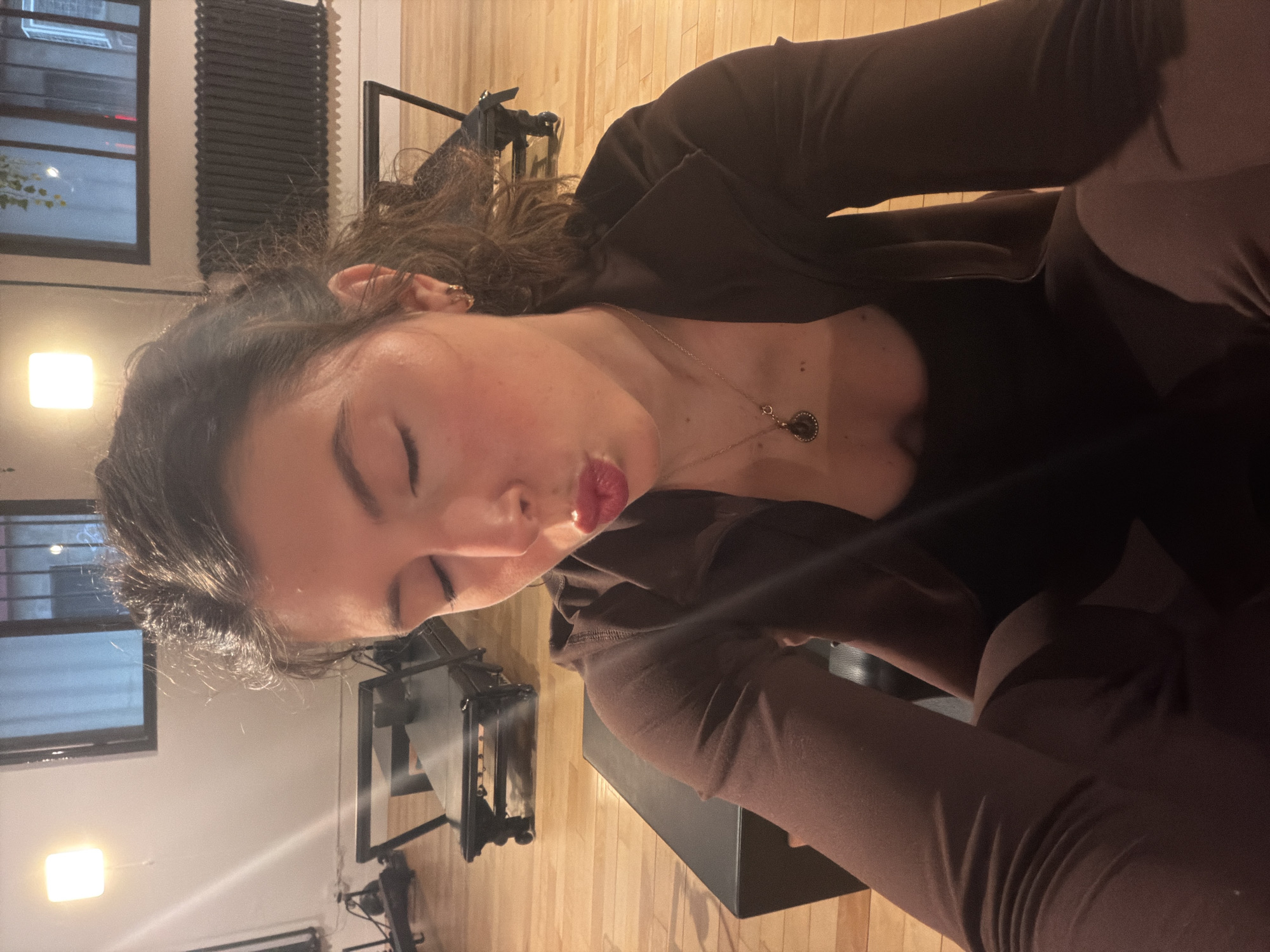 A young adult woman is captured in a close-up, rotated portrait, making a playful "kiss face" with her eyes closed and lips pursed. She has dark, wavy hair and is wearing a dark brown long-sleeved top and matching pants. A necklace with a dark, possibly metallic, circular pendant hangs around her neck. Her skin appears fair with a hint of blush on her cheeks, and her lips are colored a reddish hue.
The setting is an indoor space, likely a studio or a spacious room, characterized by a light-colored wooden floor and light-toned walls. In the background, a large window with multiple panes reveals an urban exterior with some ambient light, suggesting it is evening or nighttime. Adjacent to the window, a dark, vertically ribbed structure, possibly a radiator or window blinds, is visible. Artificial lighting is provided by a bright square light fixture on the wall in the lower left portion of the image (when oriented correctly). Dark-colored exercise equipment, possibly a Pilates reformer, can be seen in the mid-ground and background, further indicating a studio environment. No visible text is present in the image. The location is Montreal, Canada.