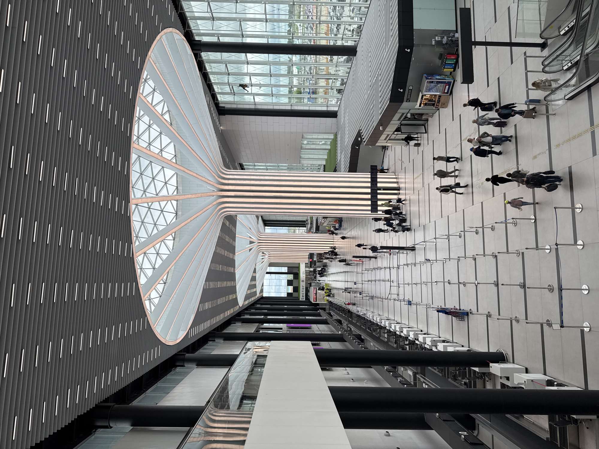 The image shows an interior view of Novosibirsk Tolmachevo Airport in Ob, Russia. From an elevated perspective, the scene captures a modern architectural design featuring a large oval skylight that extends down the length of the hall with vertical bands of lighting. Groups of people are walking along the tiled floor, some pulling luggage. Nearer to the right, there are queuing lanes, likely for security or check-in. Above them, a vending machine, an escalator, and signage are visible. In the lower part of the frame, the view is obstructed by architectural elements like pipes and white structures. The overall environment is well-lit.