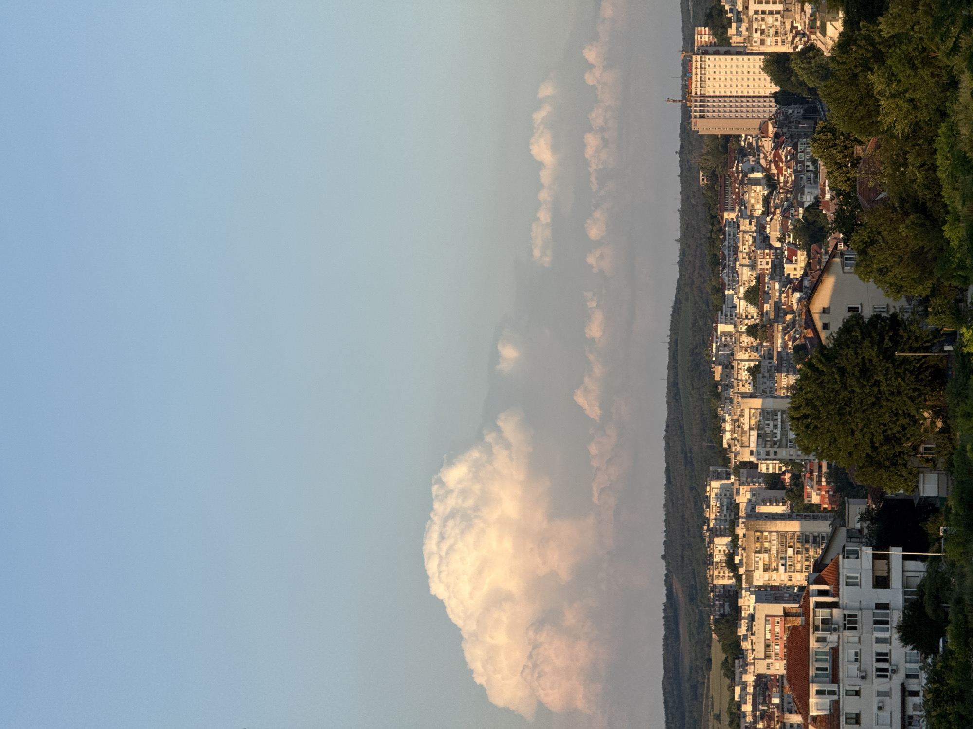 This elevated panoramic image captures a late afternoon or early evening view of the city of Pleven, Bulgaria. The sky transitions from a clear, light blue overhead to a paler, warmer blue near the horizon. A large, well-defined cumulonimbus cloud, illuminated by warm, golden sunlight on its upper contours, dominates the upper-left quadrant, while smaller, scattered cumulus clouds are visible across the mid-horizon.
The distant background consists of gently rolling hills covered in dense green vegetation. The mid-ground and foreground are filled with the urban landscape of Pleven. Numerous multi-story buildings, predominantly residential apartment blocks of varying ages and architectural styles, populate the scene. Many exhibit characteristics of post-socialist era construction, featuring concrete facades and repetitive window patterns.
A prominent landmark, the Hotel Rostov (also known as Hotel Balkan), a tall, slender, light-colored building with a grid of windows and an antenna structure on its roof, is clearly visible on the far right. Faint red lettering is discernible near the top of this building. In the lower-left foreground, a distinct white residential building with a reddish-orange tiled roof, multiple windows, and visible air conditioning units is present. Dense patches of green trees are interspersed throughout the urban environment, particularly in the foreground and between clusters of buildings. The scene is bathed in warm, golden hour sunlight, which illuminates building facades and the large cloud, while casting long shadows among the structures and foliage. No individuals or specific activities are discernible within the frame. The perspective suggests a view from an elevated point, looking north-northeast over the city center of Pleven, with the Hotel Rostov serving as a key orienting feature.