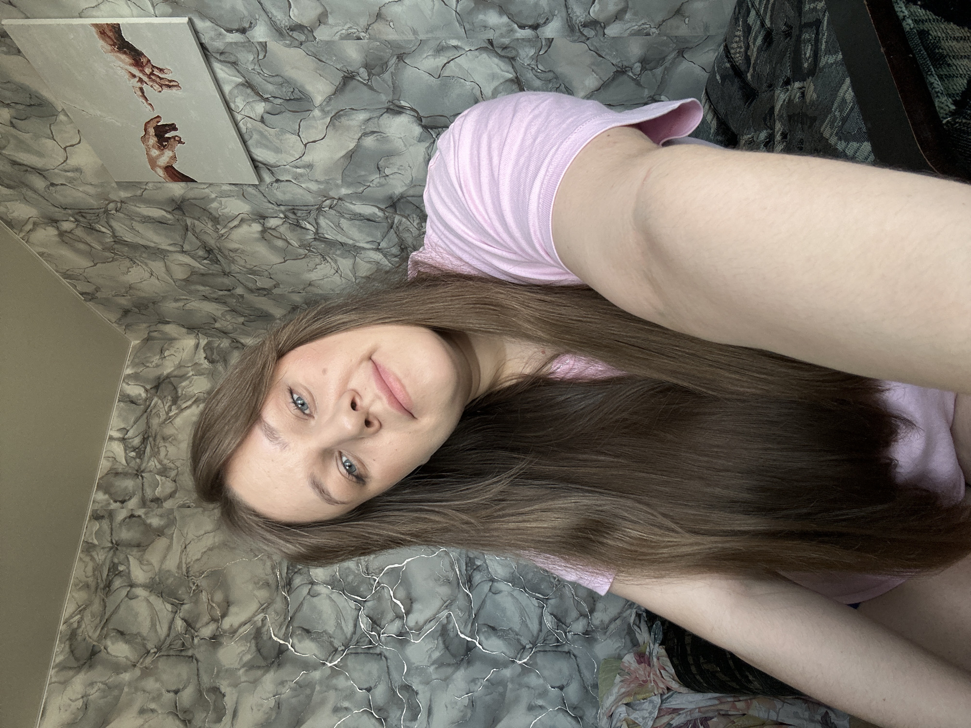 A woman with long brown hair and blue eyes is looking at the camera. She is wearing a pink t-shirt. Behind her is a gray marble patterned wall with a painting of the Creation of Adam hanging on it. A patterned dark-colored fabric is visible in the upper right corner of the image. Location: likely Ольховка, Russia.
