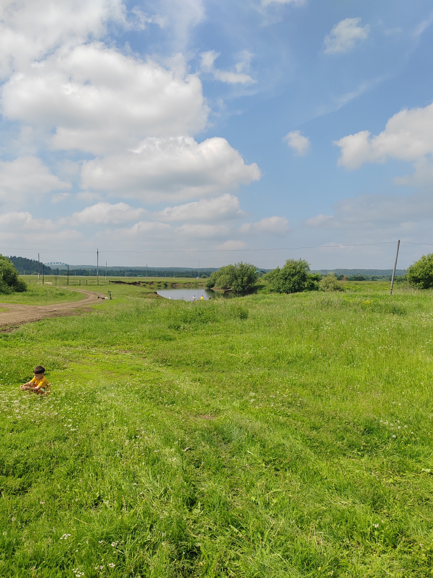 A bright, sunny day with scattered white clouds in a blue sky. A young child in a yellow shirt sits in a field of tall green grass dotted with small white flowers. In the mid-ground, a dirt path curves towards a metal gate with an archway. Beyond the path and to the right is a body of water, possibly a river or lake, with several people visible on the bank, some appearing to be fishing. The background features lush green trees and rolling hills under the expansive sky. The scene depicts a peaceful outdoor setting, likely in a rural or park area.