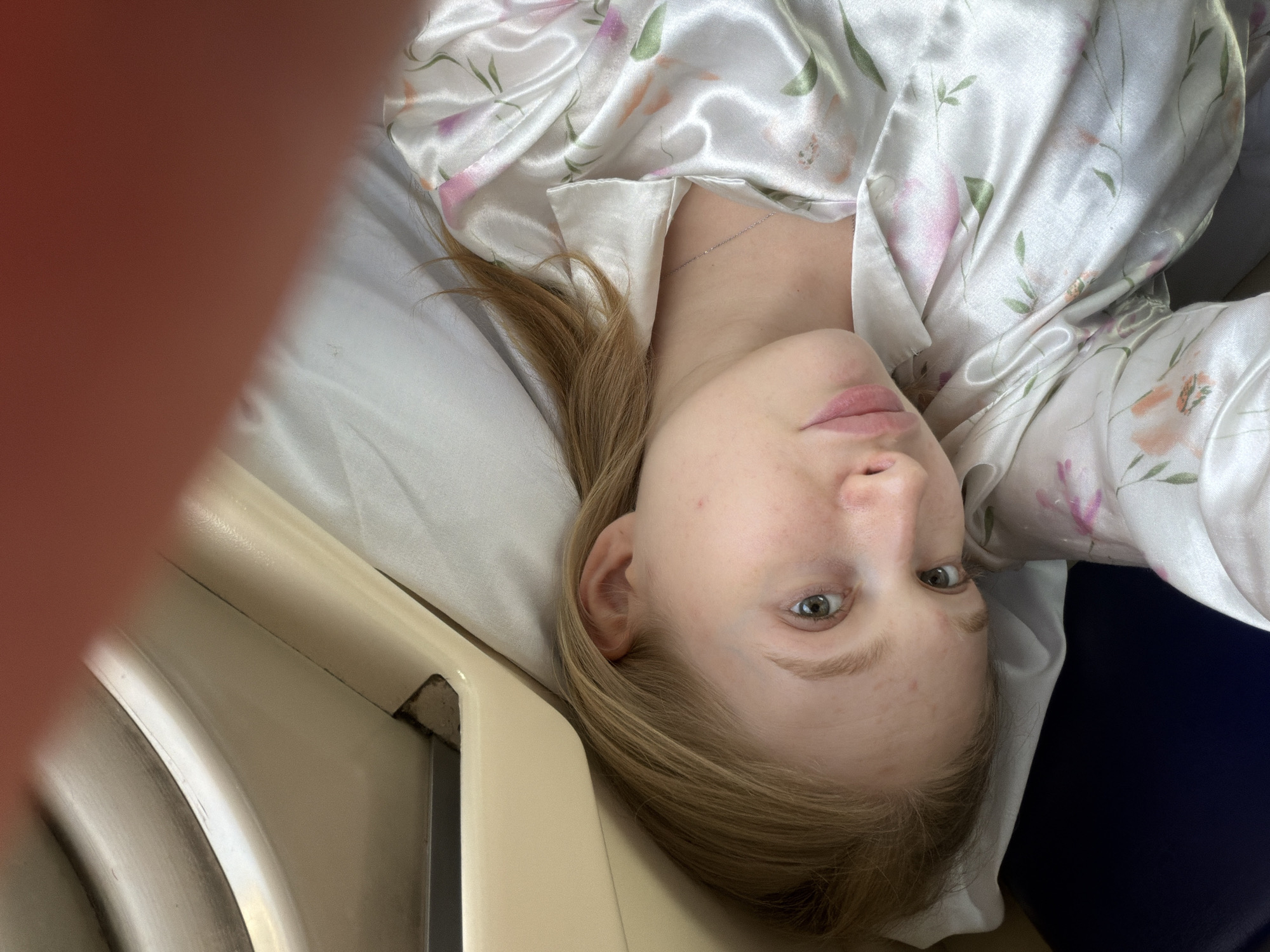 A woman is lying on a bed in what appears to be a hospital room. She is wearing a white satin robe with a floral pattern. She has blonde hair, fair skin, and blue eyes. The bed has white sheets and a light-colored side rail. Part of the image is obstructed by a blurry red object, possibly a finger. There is no indication of location.