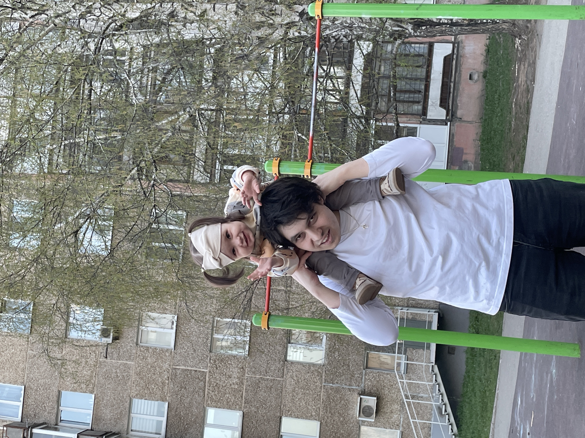 A person is holding a young child on their shoulders next to playground equipment. The playground equipment consists of a horizontal green bar with a red ladder attached. The person is wearing a white long-sleeved shirt and black pants. The child is wearing neutral-toned clothing and a white headscarf. The background includes an apartment building. The setting appears to be a playground or recreational area in Izhevsk, Russia.