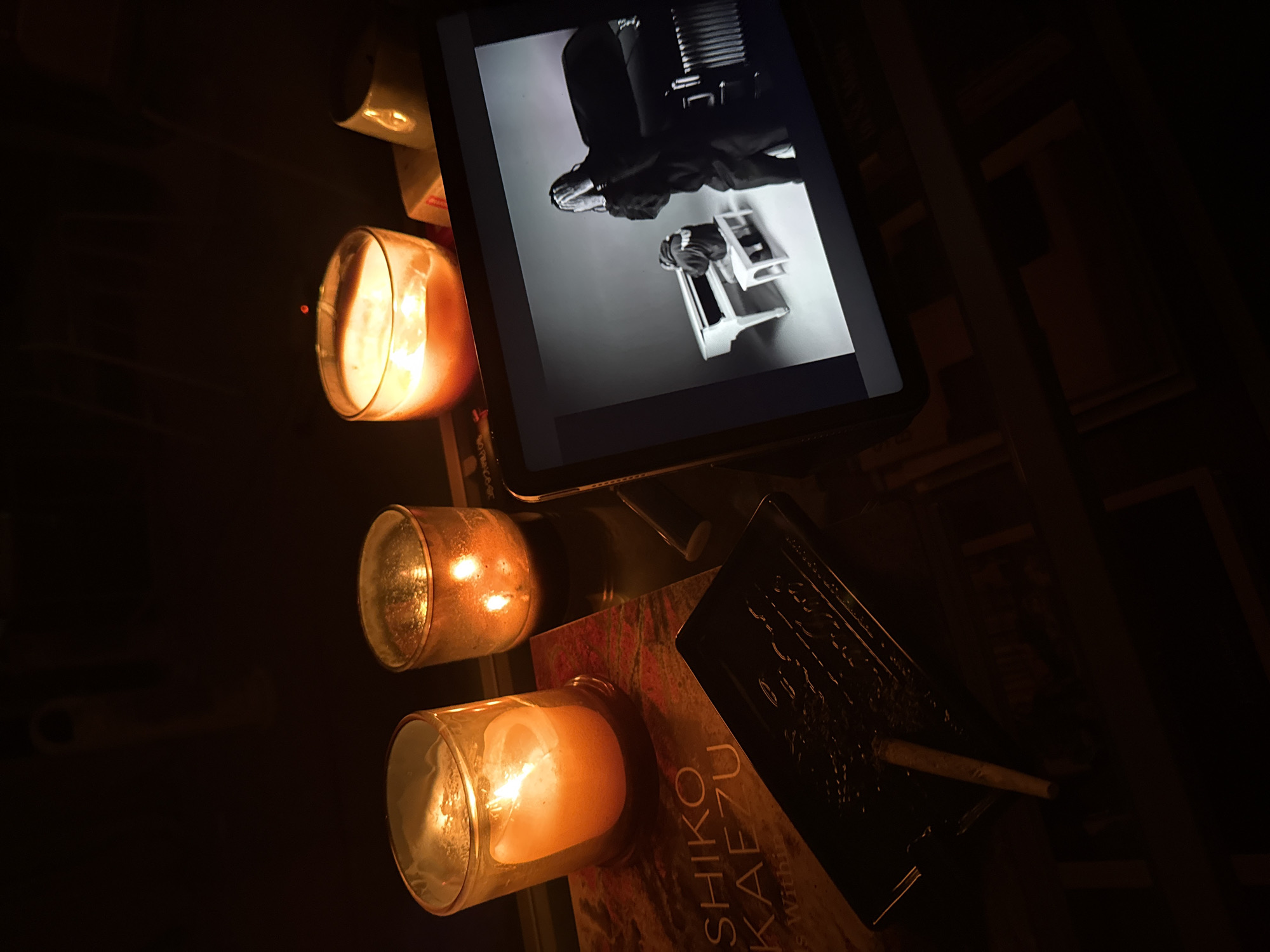 The scene is dimly lit, with three lit candles in glass holders providing the primary illumination. The candles are arranged vertically from top to bottom, with their flames casting a warm glow. A tablet displaying a black and white image is present, showing a person seated in a chair playing an accordion. Beneath the bottom candle, a book with visible text "SHIKO KAEZU" is partially visible. A dark, possibly lacquered, rectangular object rests next to the book, with a thin, light-colored object lying on top of it. The setting appears to be indoors, possibly in a room with dark furniture and shelves. There is no evidence to suggest it is in Milan, Italy.