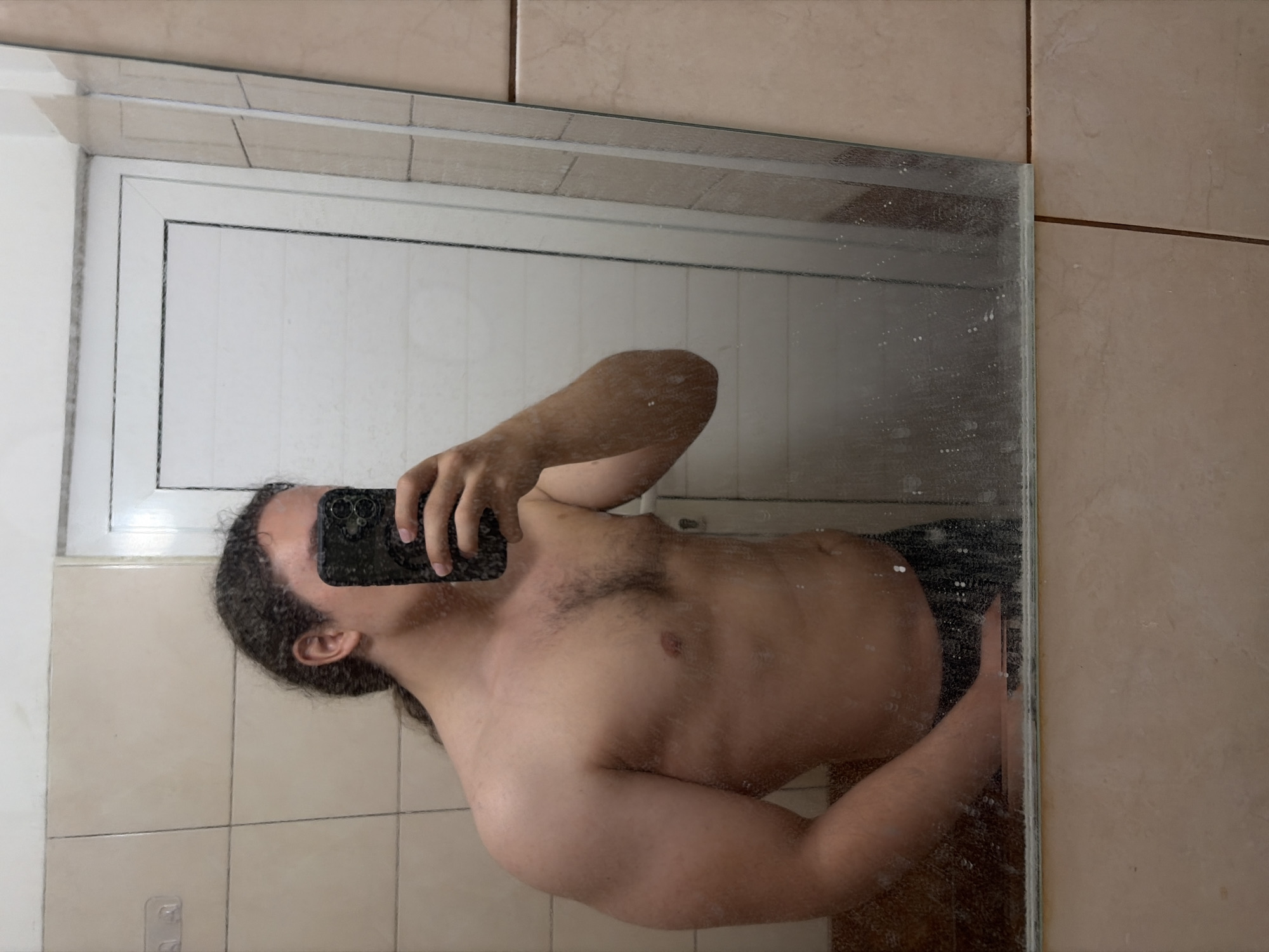 An image shows a shirtless person taking a mirror selfie, rotated 90 degrees clockwise. The person, appearing to be male, has curly dark hair and is holding a black smartphone with multiple camera lenses up to their face to capture the reflection. Their torso and right arm are visible, revealing some chest hair.
The mirror surface is noticeably dirty, covered with streaks and numerous water spots, suggesting a bathroom setting. Reflected in the mirror behind the person is a white door or cabinet, featuring a distinct dark frame outline. The walls surrounding the mirror and visible in the reflection are covered with light beige or brown tiles. One cropped image reveals a small white adhesive hook attached to the tiled wall, reflected in the mirror. The scene is indoors, likely illuminated by artificial light, making the time of day indeterminate. There is no visible text. The provided context indicates the location is Plovdiv, Bulgaria.