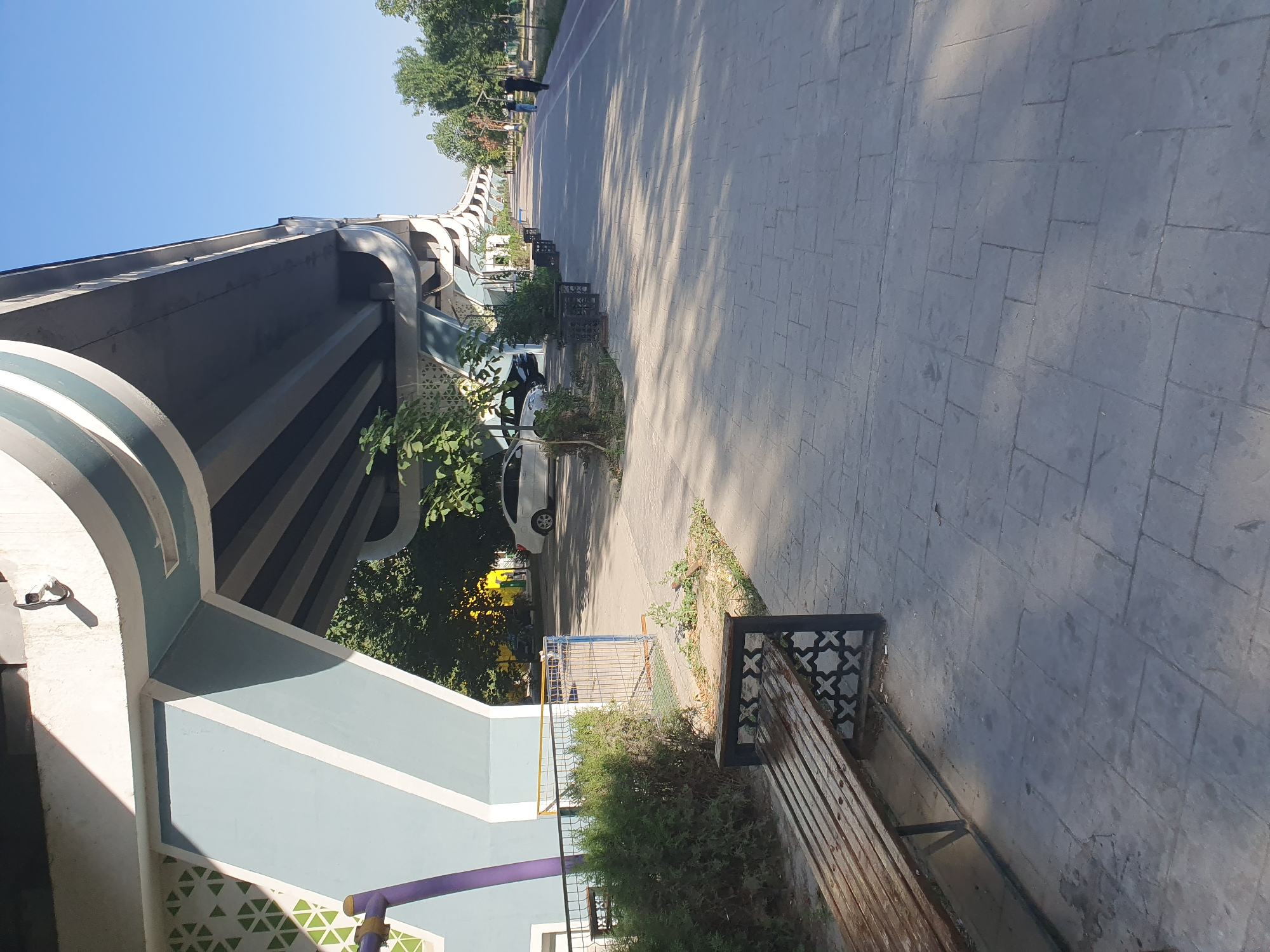 The image depicts a street scene in Tashkent, Uzbekistan, characterized by a large overpass structure with distinctive blue and white accents. In the foreground, a wooden bench with decorative black metal supports sits on a paved walkway. To the left of the bench, there is a manicured hedge and a purple pole. Beyond the hedge, a white car and a dark-colored vehicle are parked on a street that runs parallel to the walkway.
The walkway is paved with rectangular grey stones and is lined with trees and planters with decorative black metal borders. Further down the walkway, several people are visible walking, including two individuals in the distance on the right side of the image, one in a black shirt and the other in a blue shirt. The sky above is clear and blue, indicating a sunny day. The overpass itself is a prominent feature, casting shadows on the walkway. The overall impression is of an urban environment designed for pedestrian activity, possibly a park or a recreational area beneath the overpass.