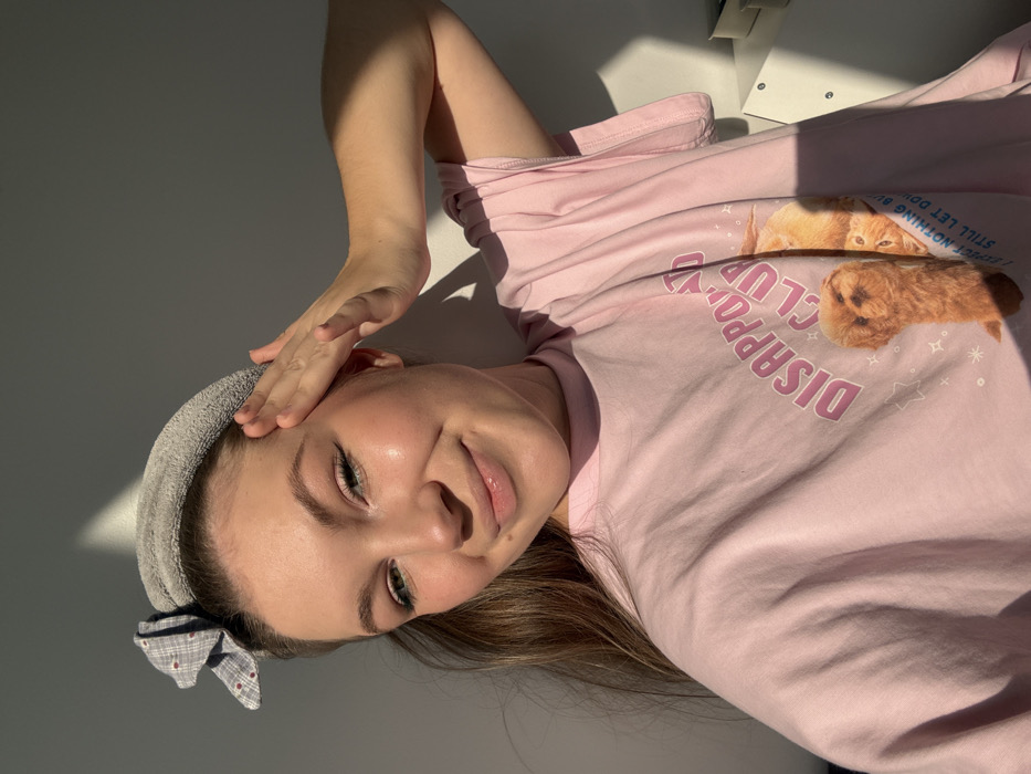 A female individual is depicted in a close-up, chest-up portrait, positioned indoors within the city of Vozdovac Urban Municipality, Serbia. She has fair skin, light brown hair partially pulled back by a grey, plush headband adorned with a grey and white checkered bow featuring small red dots on the viewer's left. Her left hand is raised, fingers slightly spread, with the palm resting on her left temple area. She wears light eye makeup and lip gloss, exhibiting a gentle smile while looking directly into the camera. She is dressed in a short-sleeved pink T-shirt, which displays a graphic that reads "DISAPPOINTED CLUB" in pink text. Below this, images of two animals are visible: a light brown, fluffy canine-like creature standing upright, and a partially obscured orange feline-like creature appearing inverted. The background is a plain light-colored wall. Strong, directional sunlight illuminates the left side of her face and casts pronounced shadows on the right side of her body, the wall, and a light-colored, indistinct furniture item visible on the far right edge of the frame.