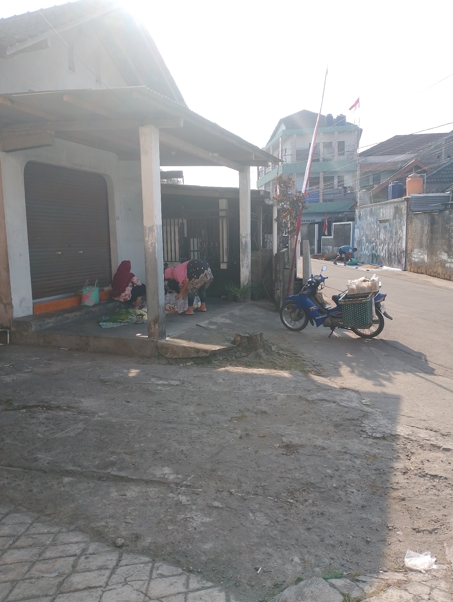 The image depicts a street scene in Tlogoadi, Indonesia, under bright daylight. In the foreground, a dusty, cracked concrete road leads away from the viewer. To the left, a low building with a corrugated metal roof extends over a sidewalk. A closed metal shutter is visible on this building. On the sidewalk, two women are engaged in activity. One woman in a red headscarf sits with her back to the camera, while another in a pink top and patterned skirt bends over, seemingly organizing items on the ground. Several plastic bags and what appear to be vegetables are scattered around them.
Further down the street, a blue Suzuki motorcycle is parked on the side of the road. It has a green woven basket attached to the rear, containing boxes and what looks like produce. Beyond the motorcycle, a man in a blue t-shirt is bent over near a white tarp laid out on the ground, with bags of what might be grain or sand next to him.
In the background, several buildings are visible. A multi-story building with green and white accents stands out, adorned with Indonesian flags. Other buildings with pitched roofs and water tanks are also present, along with power lines. A tall pole with flags is also prominent in the background. The overall environment suggests a residential or commercial area in a developing urban setting, with a mix of simple structures and more modern buildings. The sunlight casts distinct shadows across the scene, indicating it is likely morning or late afternoon.