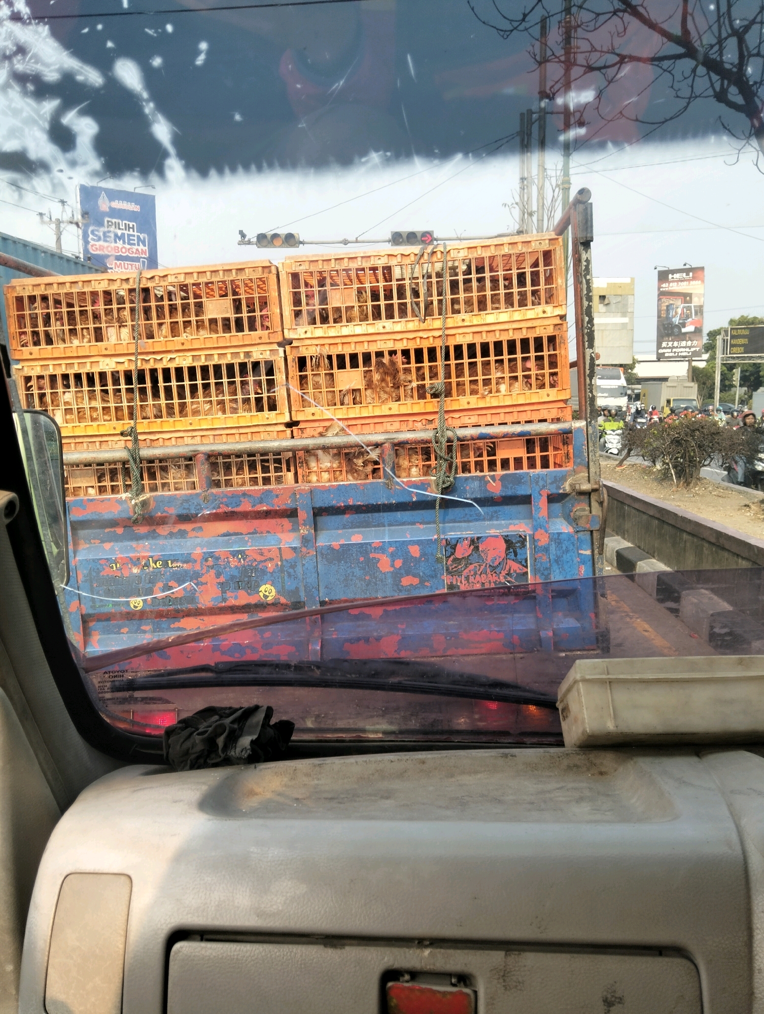 The image is a photograph taken from inside a vehicle, looking out through the windshield. The primary subject is a blue truck carrying a stack of orange crates filled with chickens. The truck's bed is weathered and shows signs of rust and peeling paint. A rope is tied to one of the crates and hangs down the side of the truck.
In the background, traffic lights are visible, suggesting the truck is stopped at an intersection. Various signs are present, including a blue billboard with Indonesian text that appears to advertise cement. Another billboard in the distance shows an image of a forklift and has text in a foreign language, possibly Chinese. Other vehicles, including motorcycles and cars, are also present in the background, indicating a busy urban environment. The overall scene suggests a street in Semarang, Indonesia, likely during daylight hours. The interior of the vehicle from which the photo is taken is partially visible, showing the dashboard and a black cloth resting on it.