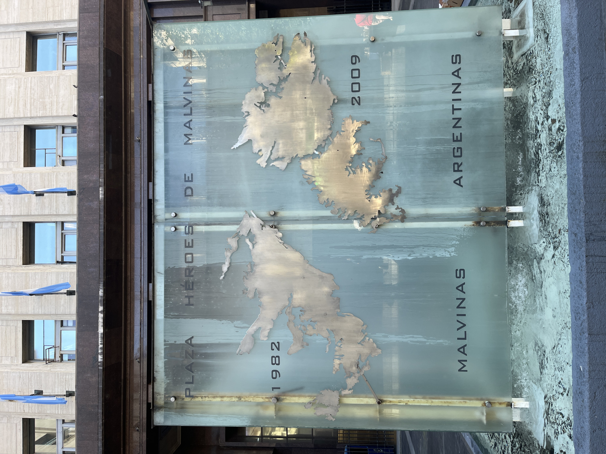 The image displays a vertically oriented public monument, likely located in Buenos Aires, Argentina. The central feature is a large, frosted glass panel, which serves as a canvas for metallic cutouts and inscribed text.
Affixed to the glass are two distinct metallic shapes representing the islands of the Malvinas (Falkland Islands). The lower map is accompanied by the horizontal year "1982," while the upper map is associated with the year "2009."
Text is etched onto the glass panel, reading vertically from bottom to top. On the left side, it says "PLAZA HÉROES DE MALVINAS." On the right side, it reads "MALVINAS ARGENTINAS."
The background on the left reveals a tall building with a light-colored, textured facade and multiple rectangular windows. From some of these windows, blue and white ribbons, emblematic of the Argentine flag, are hanging. A dark, possibly granite or stone, column or wall separates the building from the monument. At the bottom right, a water feature, perhaps a fountain or reflecting pool, is visible, showing some green algae or moss, suggesting an outdoor setting exposed to elements. The glass panel itself appears wet or streaky with watermarks.
The monument commemorates the Falklands War (Guerra de Malvinas) of 1982, honoring "heroes" and asserting the Argentine claim over the islands, possibly referencing an event or declaration in 2009. The scene is brightly lit, indicating daytime.