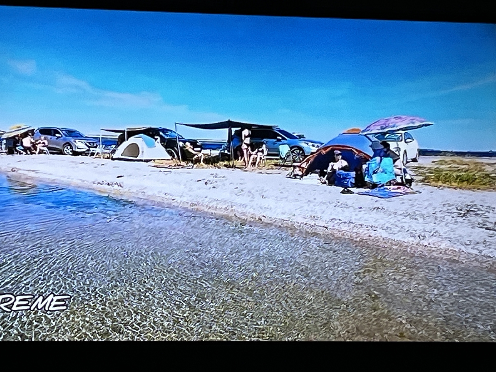 The image shows a beach scene with several tents and vehicles set up along the shore. The water in the foreground appears clear and shallow, with sunlight reflecting off the surface. In the background, there are people gathered around the tents and vehicles, some sitting in chairs and others standing. The sky is a clear blue, indicating a sunny day. There are multiple cars, including a silver SUV, and a white car. Tents and awnings are set up to provide shade. Two tents are visible, one white and dome-shaped, and another larger one with blue and orange fabric. There is a patterned umbrella shielding a group of people. The ground is sandy, with some sparse vegetation in the distance. The word "XREME" is visible in large white letters in the lower left corner of the image, superimposed on the water. This appears to be a recreational camping or beach gathering. The location is not identifiable as being in Kharkiv, Ukraine.