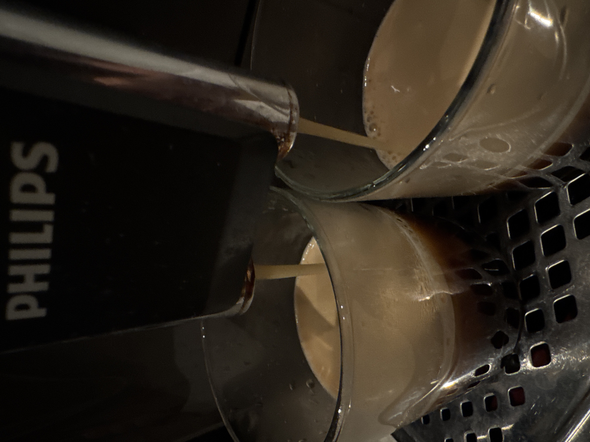 In a close-up, rotated image, a Philips coffee machine dispenses a light brown beverage, likely coffee with milk, into two clear glass cups. The machine's dark body, bearing the vertically oriented "PHILIPS" logo in white, is visible on the left side of the frame. Two metallic spouts extend from the machine, with streams of the frothy, milky liquid pouring into the cups.
The liquid, which appears to be forming a layered or frothy texture, is partially filling both transparent glass cups. The cups rest on a dark, metallic grid or drainer with square perforations, suggesting a coffee machine's drip tray. The scene is illuminated by artificial light, creating reflections on the glass and metal surfaces and highlighting the texture of the drink. The overall lighting is somewhat dim, contributing to a cozy or intimate atmosphere. The perspective is from a slightly low angle, focusing on the dispensing action and the filling cups. No people are visible, and the setting appears to be an indoor environment, possibly a kitchen counter.