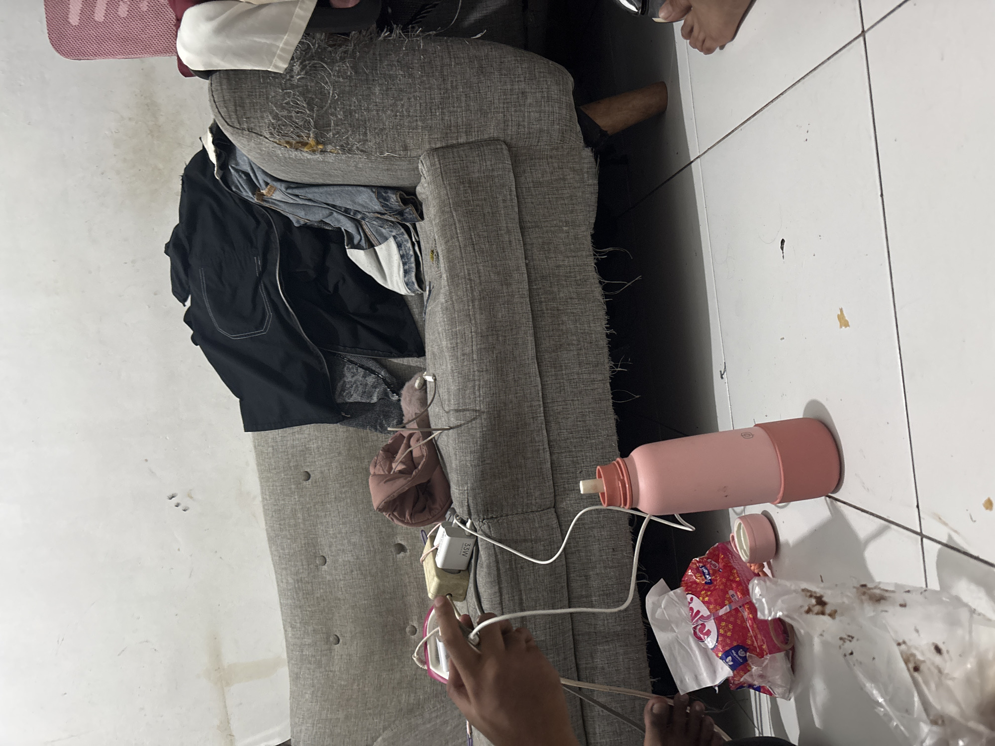 A cluttered living room in North Jakarta, Indonesia, features a worn grey sofa with clothing piled on top, including a black jacket and denim jeans. Scattered around the sofa are various items: a pink water bottle, a pack of tissues, a small pink container, and some plastic-wrapped food. Several electrical chargers and cords are also visible on the sofa and extending to the floor, suggesting electronic devices are being charged. The walls are stained and discolored, indicating a lack of recent maintenance. The floor is tiled and has some scattered debris. A person's hands are partially visible, interacting with the charging cords. Another person's feet are visible on the floor in the lower right corner of the image.