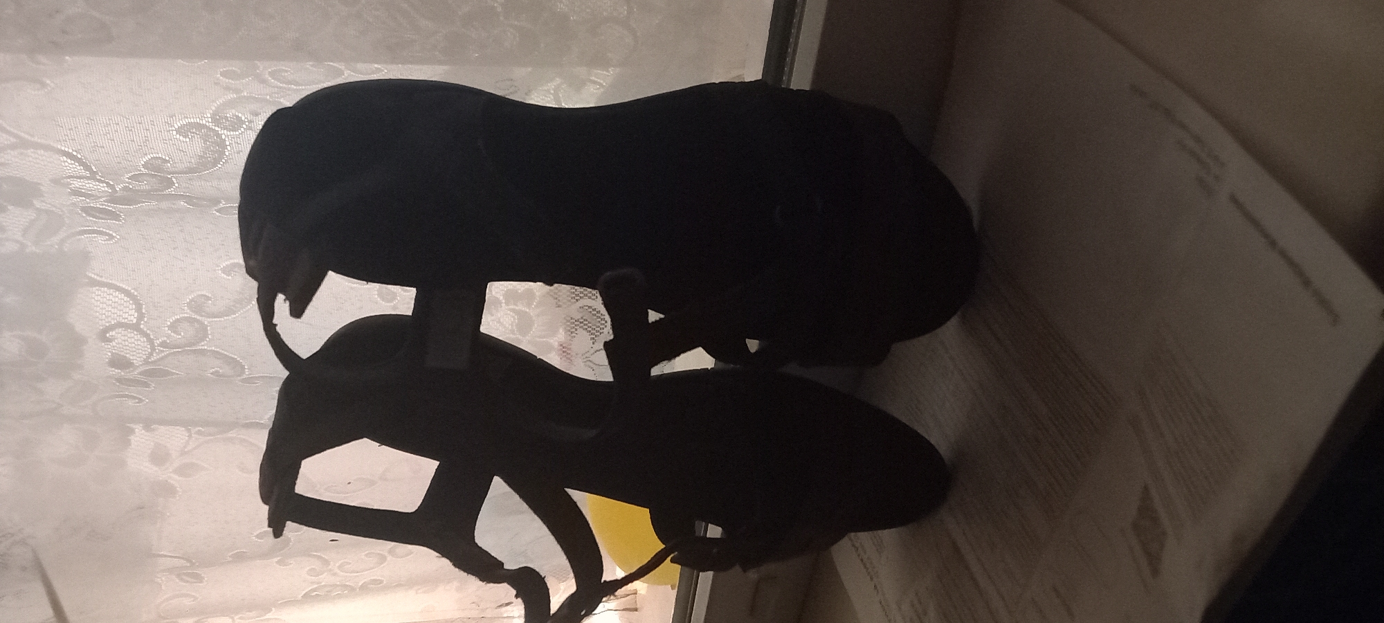 In Kryvyi Rih, Ukraine, a pair of black high-heeled sandals are silhouetted against a window with a lace curtain. The sandals, with straps and open toes, are positioned on a windowsill. Below the windowsill, a document with visible text lies on a surface. The scene is dimly lit, with the light source appearing to be behind the window.