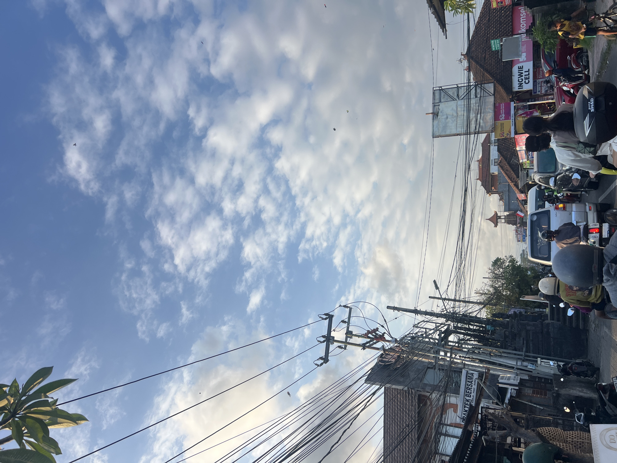 The image depicts a street scene in Denpasar, Indonesia, likely during daylight given the bright sky. The foreground is dominated by a traffic jam, with numerous motorcycles and a few cars visible. Several people are on motorcycles, some wearing helmets. There are buildings on both sides of the street, featuring a mix of traditional and modern architecture, with many power lines crisscrossing overhead. Signs in Indonesian are visible on some of the buildings, advertising businesses like "PRAKTEK BERSAMA" and "INGWIE CELL". The sky is blue with scattered white clouds, and there appear to be a few kites flying high above. The overall impression is of a busy urban environment with a blend of cultural elements.