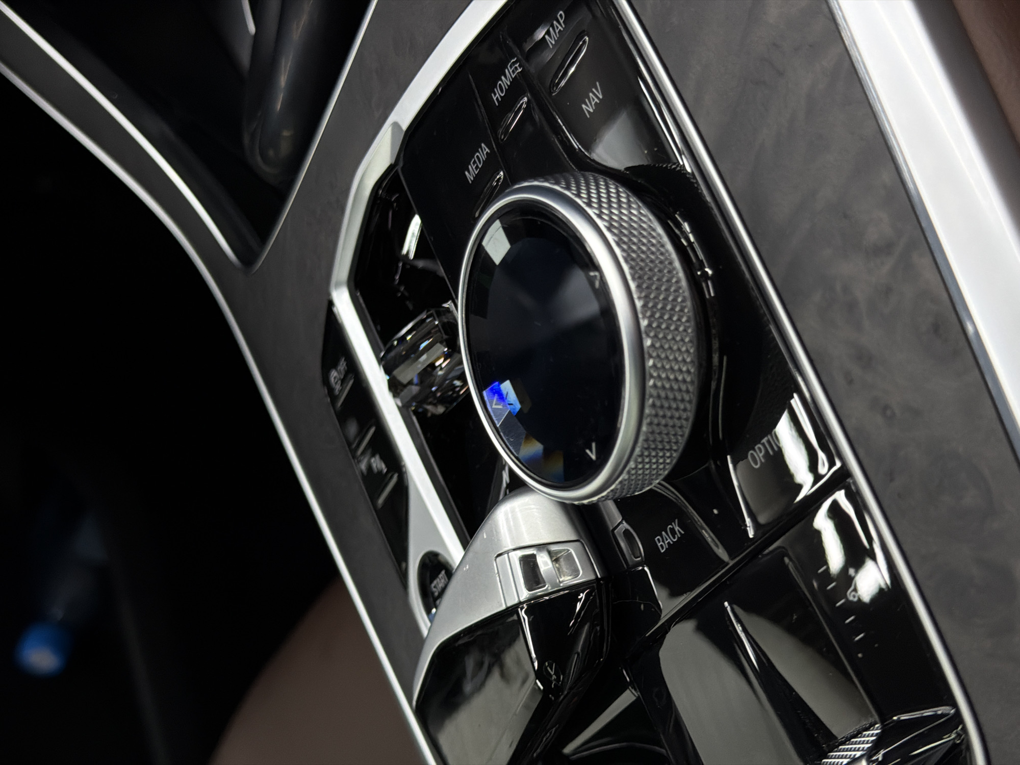 The image shows the center console of a BMW automobile. The console features buttons labeled "HOME," "MEDIA," "MAP," "NAV," "OPTION," and "BACK." A large rotary dial dominates the center, alongside a gear shifter. The trim appears to be dark wood grain. The location is not discernible from the image.