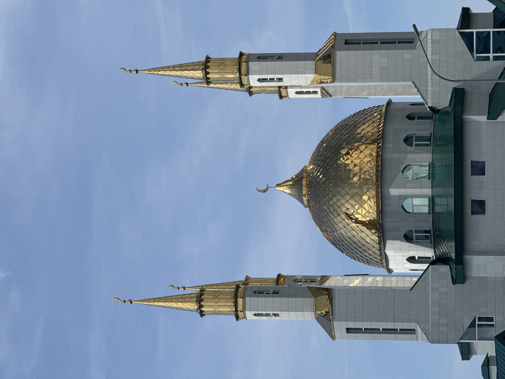 This image captures a modern mosque under a clear, bright blue sky, presumably located in Nizhnevartovsk, Russia. The central feature of the mosque is a large, shimmering golden dome, constructed from numerous faceted panels that reflect the sunlight. This main dome, and smaller spires on the structure, are each topped with a distinctive golden crescent moon and star symbol, characteristic of Islamic architecture.
Flanking the main dome are two tall, slender minarets, also adorned with golden spires that mirror the design of the dome. These minarets, along with the main body of the mosque, are constructed from light grey or white paneling, giving the building a sleek and contemporary aesthetic. Several windows, both rectangular and arched, are visible across the building's facade. Darker green architectural elements, possibly roofing or decorative cladding, provide a subtle contrast to the grey and gold.
The photograph is taken during the day, indicated by the well-lit building and the vibrant blue sky with only a few faint, wispy clouds. There is no visible human activity or text in the image; the focus is entirely on the grandeur and architectural details of the mosque.