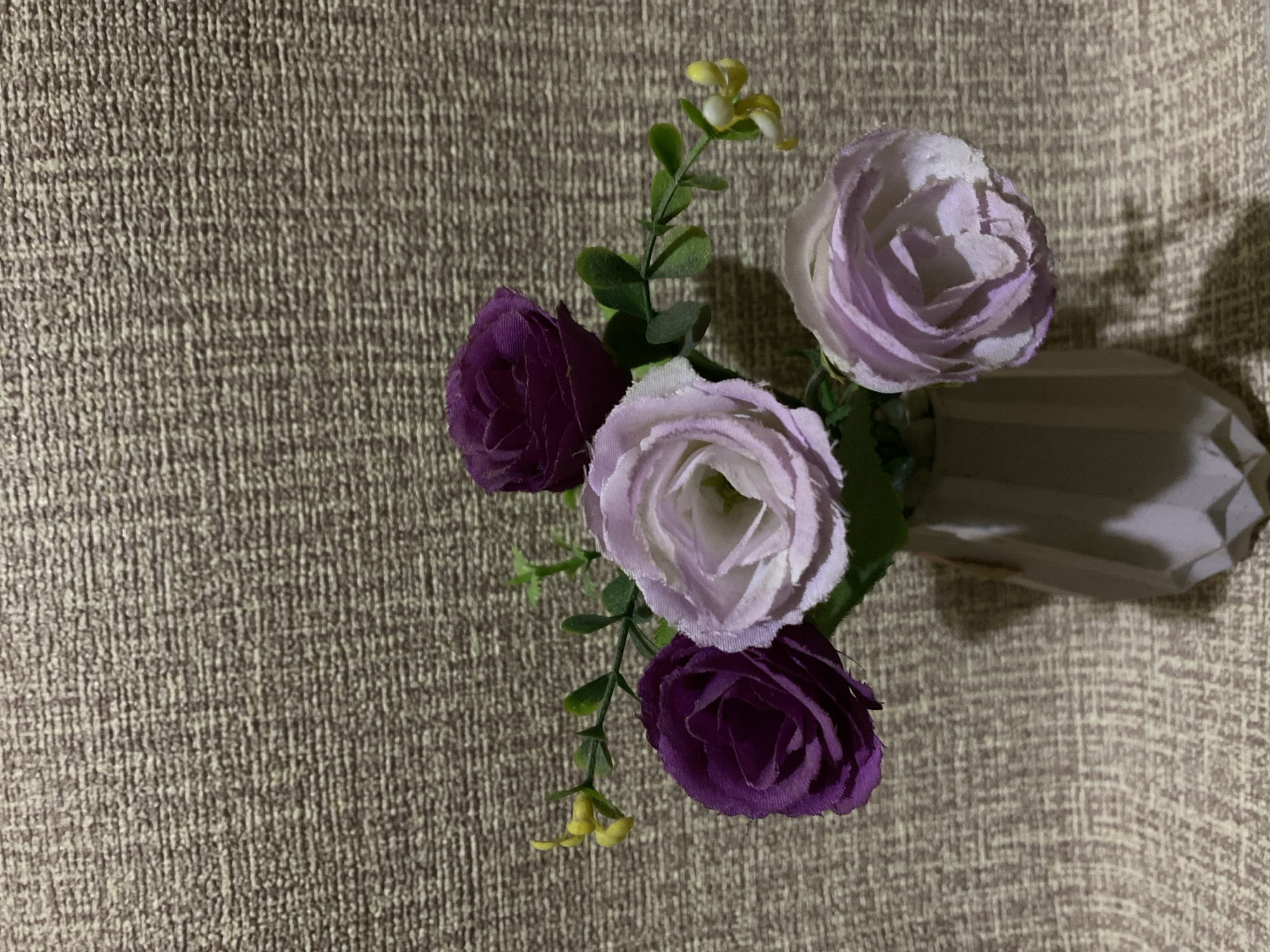 A vase containing four artificial flowers, two dark purple and two light purple, is placed on a textured, light brown background. The vase is light gray and polygonal in shape. The flowers have green leaves and small yellow buds. The lighting in the scene casts a shadow to the lower right of the vase. This image does not depict any location within the city of Gumelar Kidul, Indonesia.