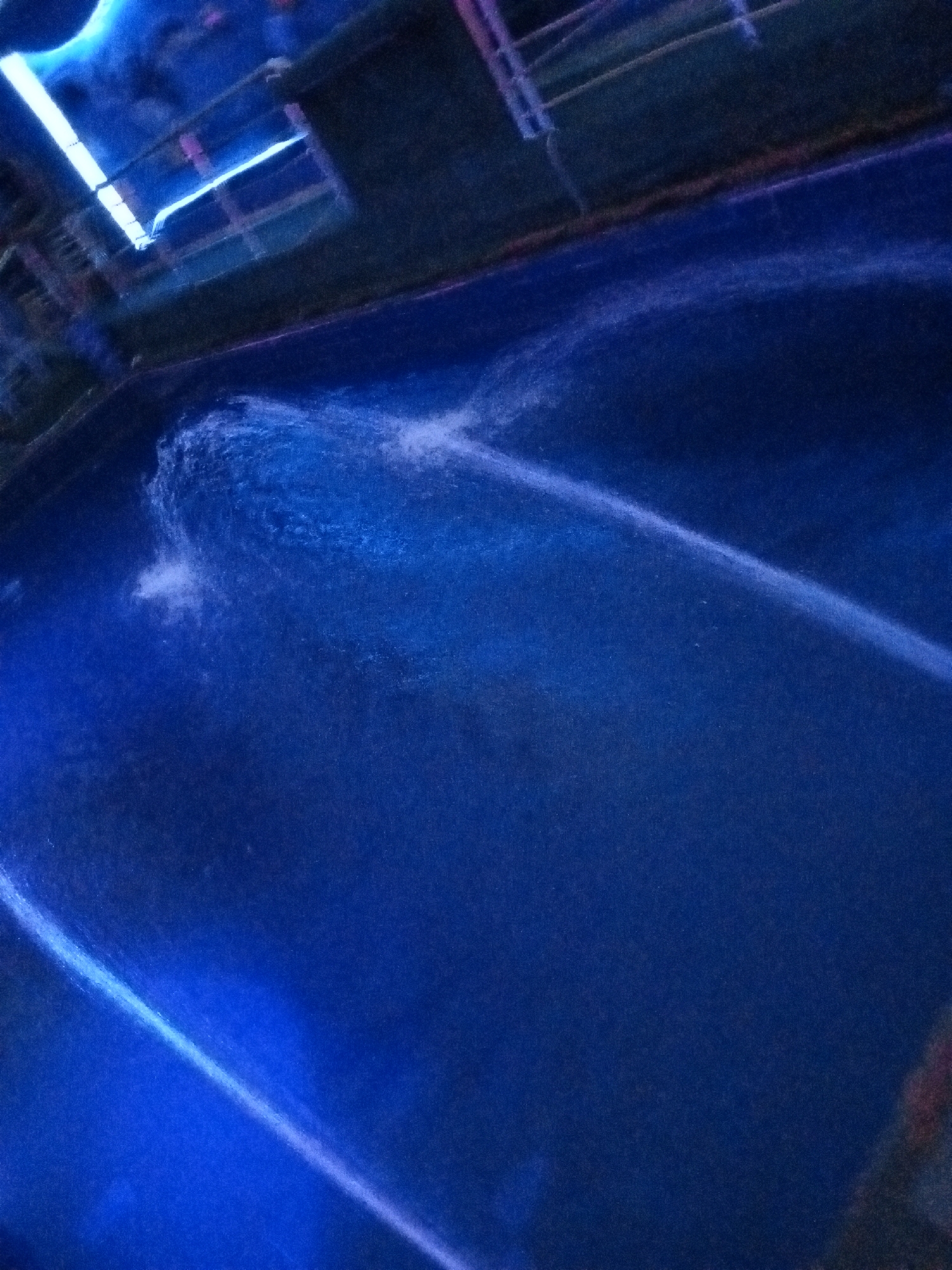 An overhead, angled shot captures a dark blue pool of water. In the top left corner, a bright blue neon light strip is visible above the pool, casting a reflection on the water's surface. To the right of this light, a dark, structured area with railings and what appears to be seating can be seen, likely part of a pool deck or elevated platform. A thin, arcing stream of white water sprays across the surface of the pool from the upper middle towards the bottom right, creating a blurred, dynamic effect on the otherwise still water. The overall scene is dimly lit, with the blue illumination from the neon light dominating the visual. There are no people or animals present in the frame. The location appears to be an outdoor pool area.