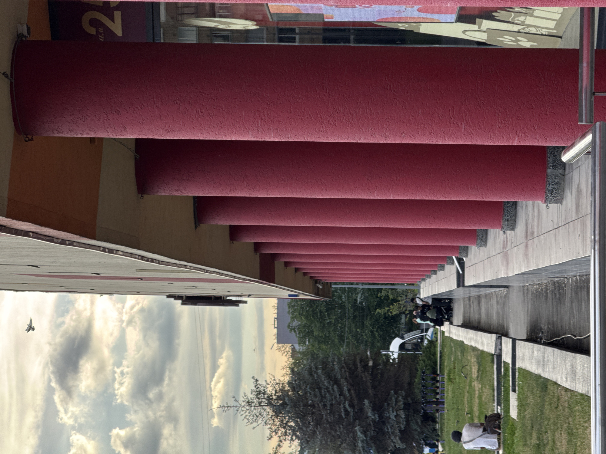 An outdoor, eye-level, vertical medium shot captures a building with red cylindrical columns in the foreground and a cloudy sky with a distant airplane. The building, with a yellow overhang, has a row of red columns that extend into the distance. To the left of the columns, a paved walkway runs alongside a grassy area. A person in a white shirt is seated on the grass with their back to the camera, accompanied by a bag. Further into the scene, a group of people are gathered near a white van. Beyond the grassy area, trees and buildings are visible. A large, dark green coniferous tree stands prominently on the left. The sky is filled with dramatic, illuminated clouds, suggesting either sunrise or sunset. The overall scene appears to be in an urban environment.