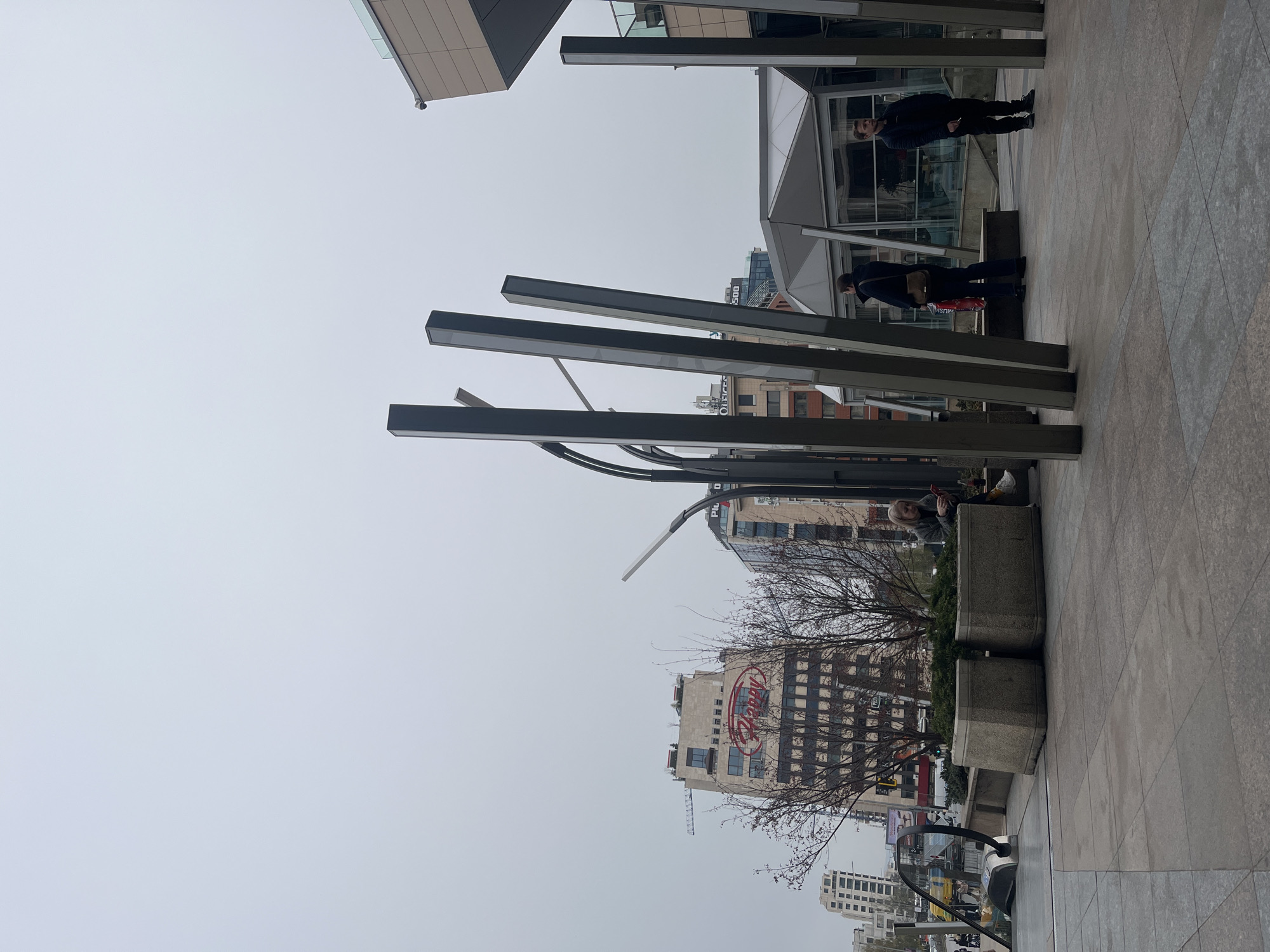 The image captures an urban scene in Sofia, Bulgaria. The foreground features a paved pedestrian area with tall, rectangular metal structures aligned vertically. Two individuals are standing near a building with glass windows and modern architectural features. A woman sits on a concrete planter further down the walkway. A bare tree stands between the planter and the background buildings. In the background, there are various buildings, one prominently displaying the word "Happy" in large letters. A construction crane is visible near the "Happy" building. Other structures and street-level activity, including cars, add depth to the urban context. The sky is overcast.