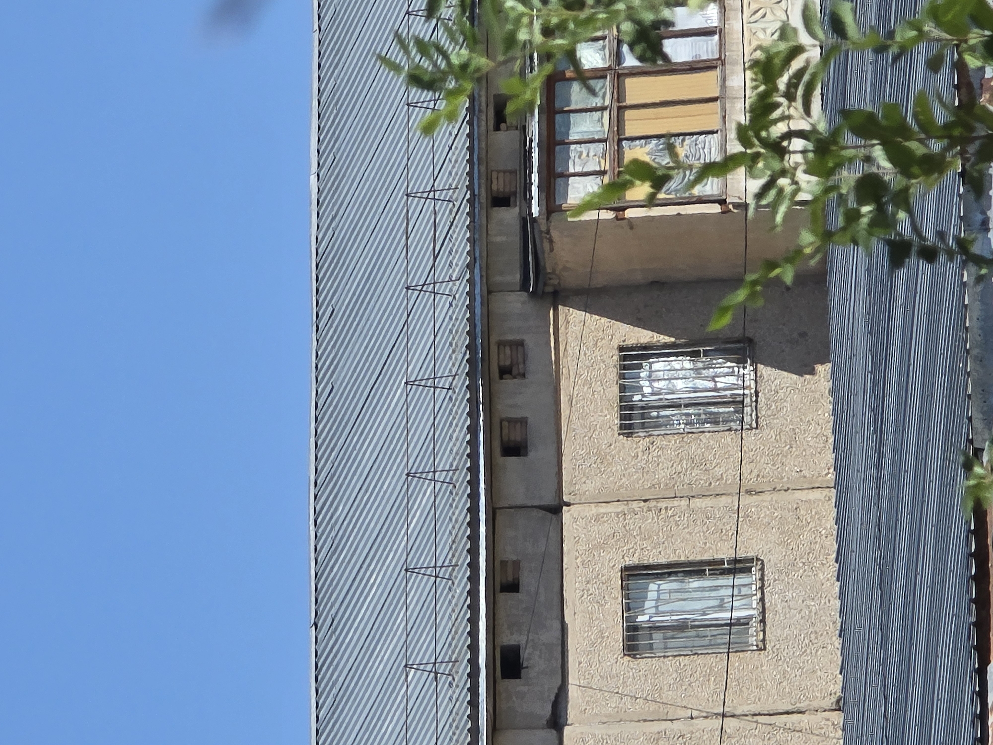 This is a daylight, eye-level, medium shot of the side of a multi-story building, with a clear blue sky in the background. The building has a corrugated metal roof, and below it, a beige-colored facade. There are several windows visible; two on the lower level with metal bars and curtains, and a balcony with a window on the upper level. The building appears to be in a state of disrepair, with some broken window panes and exposed building materials. The image is taken in Qaratau, Kazakhstan.