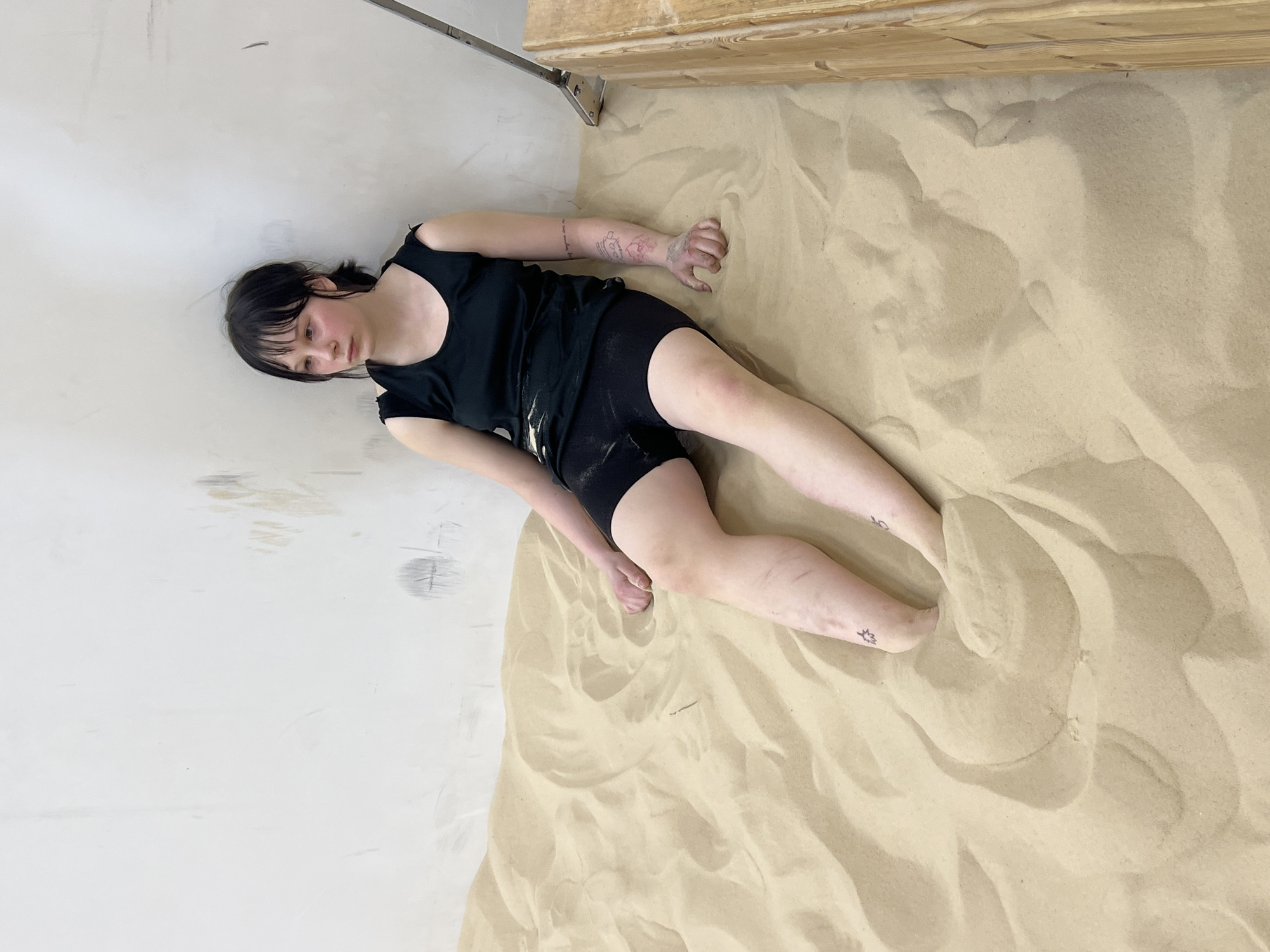 A young person with short, dark hair and bangs lies on their back in a large indoor area, partially submerged in light beige sand. They wear a black sleeveless top with cutouts on the shoulders and black shorts, both lightly dusted with sand. The person's head is turned slightly to the left, displaying a neutral, possibly contemplative, expression. Visible tattoos include script on their left forearm and abstract designs on their left upper arm and both ankles.
The setting is an interior space, likely an art installation or studio in Saint Petersburg, Russia. A plain, off-white wall with some marks and imperfections forms the backdrop on the left. The right side of the frame is dominated by the sand, which is arranged in soft, organic ripples and mounds, suggesting recent disturbance. In the upper right corner, a light-colored wooden structure with metal brackets is visible, from which a thin metal bar extends across the top of the white wall. The image is taken from an elevated perspective, looking down onto the scene, creating a quiet and introspective mood.