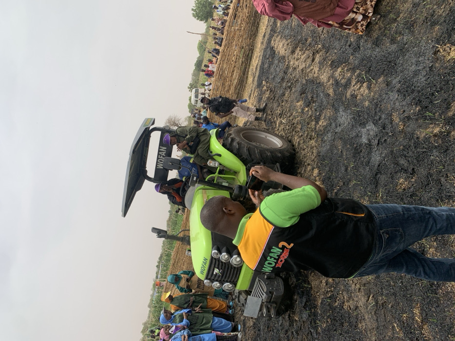 The image shows a green tractor in a field with burned vegetation. The tractor has a black canopy with the word "WOFAN" written on it. Three people are sitting on the tractor. A man in a green vest with the words "WOFAN ICON 2" printed on it is standing in front of the tractor and taking a picture with a smartphone. There are many people standing behind the tractor. The image was taken in Ringimawa, Nigeria (latitude: 11.666, longitude: 8.414). The field has patches of burnt vegetation. A white vehicle is visible in the background.