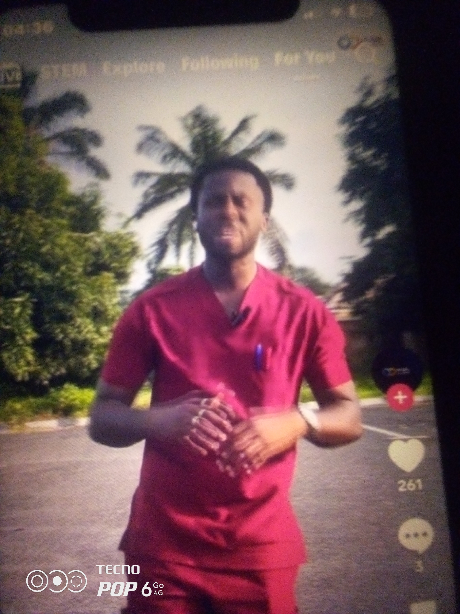 A man in a maroon scrub suit is standing outdoors, possibly in a city like Howayi, Nigeria, as suggested by palm trees and lush greenery in the background. He is looking forward and speaking, with his hands clasped in front of him. The image appears to be a screenshot from a mobile device, indicated by the timestamp "04:36" and icons for "STEM," "Explore," "Following," and "For You" at the top. The right side of the screen shows social media engagement icons, including a heart with "261" likes and a speech bubble with "3" comments. A TECNO POP 6 4G logo is visible at the bottom left. The overall scene depicts a digital content creator or presenter in a casual outdoor setting.