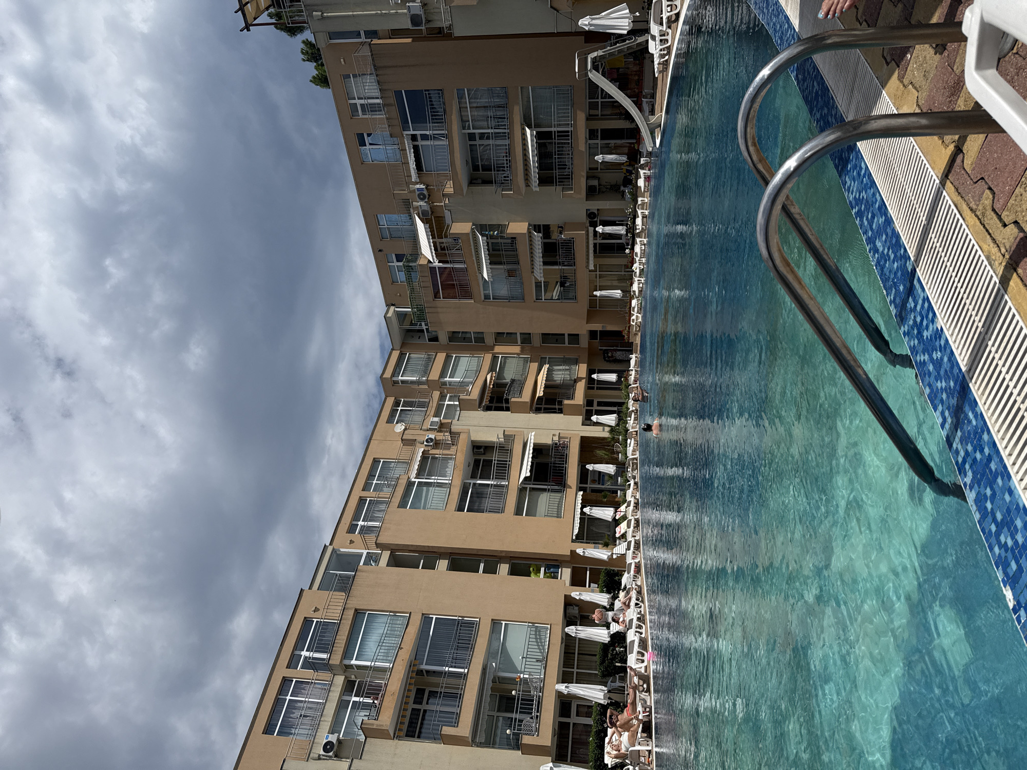 This image depicts a sunny day at a hotel pool located in Nesebar, Bulgaria. The scene is dominated by a large, turquoise swimming pool in the foreground, with its steps and metallic railings visible. Around the pool, numerous white lounge chairs and umbrellas are set up on a paved area, some occupied by people sunbathing. In the background, a multi-story hotel building with beige-colored walls and numerous balconies stretches across the frame, under a cloudy sky. A water slide is visible to the right of the pool.