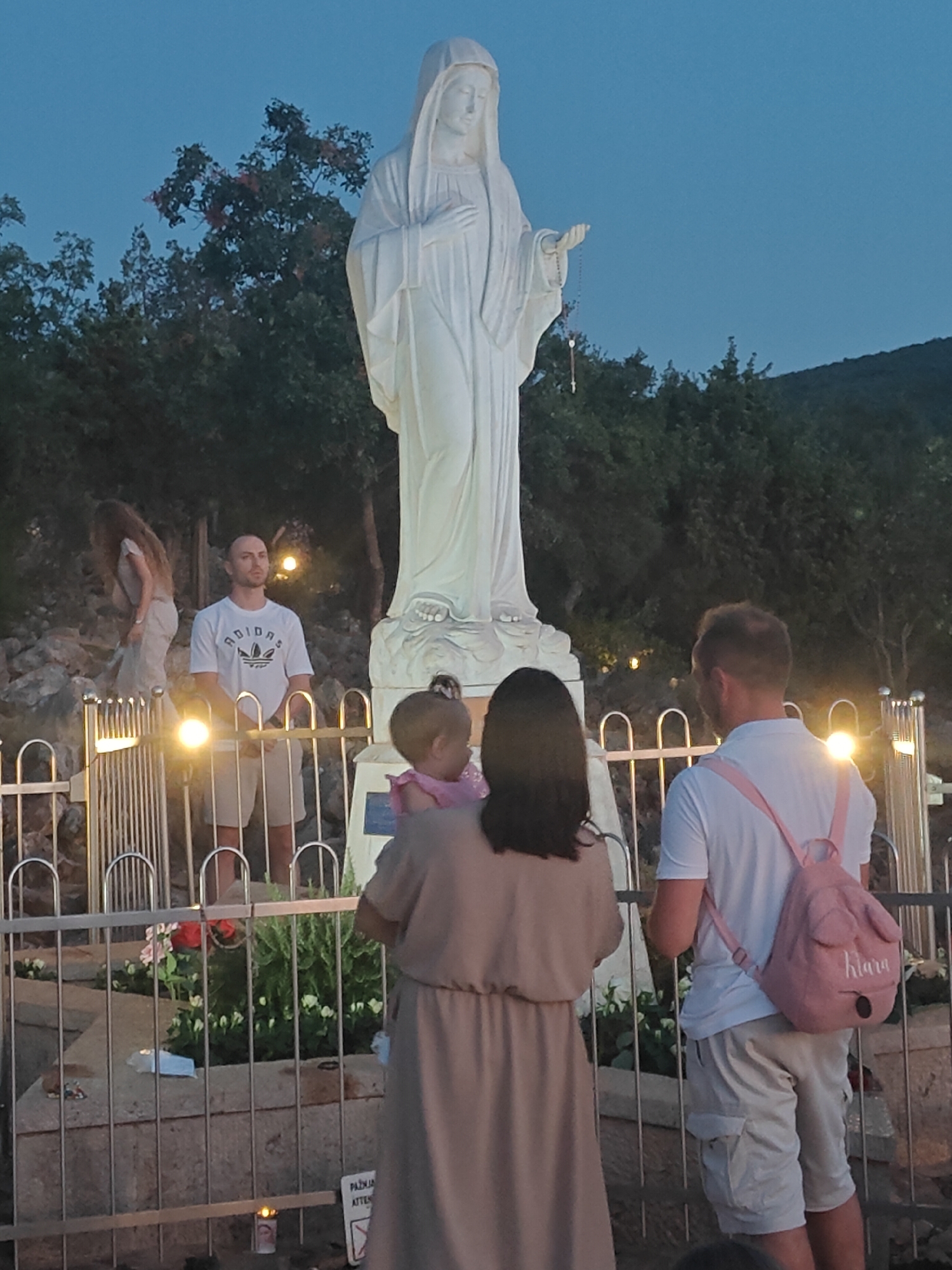 The image depicts a gathering at Apparition Hill in Međugorje, Bosnia and Herzegovina, at dusk. Dominating the scene is a large, white statue of the Virgin Mary, holding a rosary. In the foreground, a family is present: a man in a white t-shirt and beige shorts, a woman in a beige dress holding a toddler, and another man in a white t-shirt and beige cargo shorts, wearing a pink backpack with the name "Kiara" visible. The area is cordoned off by a metal railing, with decorative plants and flowers visible behind it. Several lit candles and a sign with Croatian text, possibly indicating "Attention," are also present. The background consists of trees and a hillside under a twilight sky.