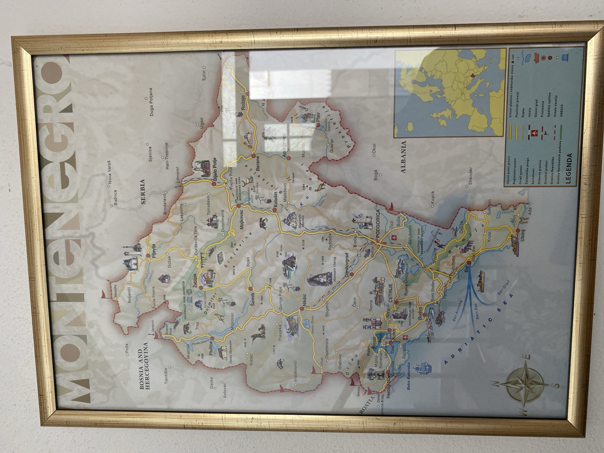 The image shows a framed map of Montenegro hanging on a wall. The map is colorful, with distinct regions, cities, mountains, and bodies of water marked. Key landmarks and national parks are also depicted with small illustrations. A legend at the bottom right explains the map's symbols, including roads, railways, airports, borders, and other geographical features. A small inset map of Europe shows Montenegro's location highlighted in red. The city of Sutomore is marked on the map along the Adriatic Sea coast, south of Bar. The overall scene is a well-lit interior shot of the map displayed on a plain wall.