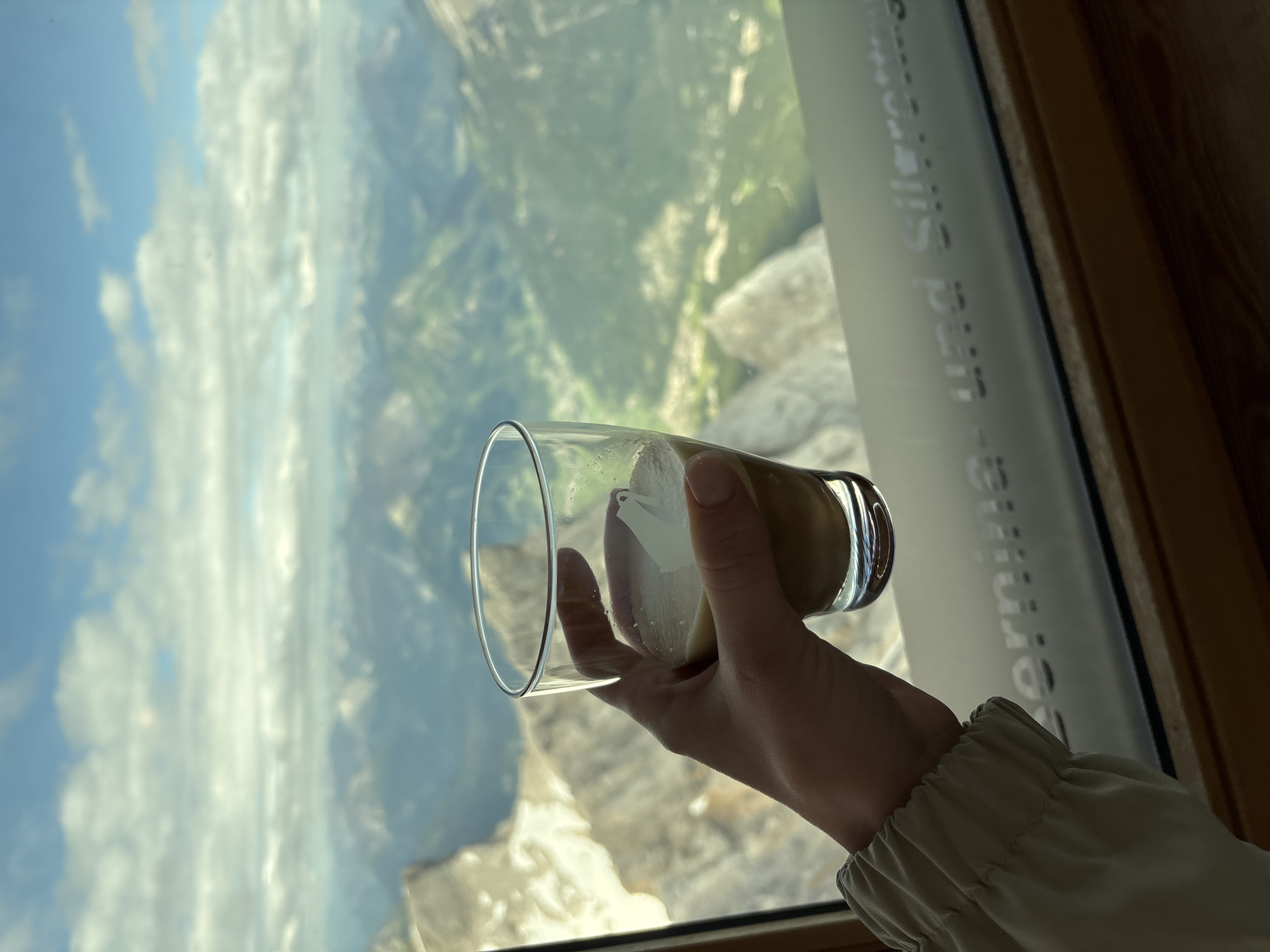 A hand holds a glass of coffee in front of a panoramic view of mountains. The coffee appears to be an espresso with foamed milk. The hand is of a person wearing a beige jacket with gathered elastic at the cuff. The background shows a vast landscape of green valleys and rocky mountain peaks under a cloudy blue sky. The image is taken from inside a building, with a window framing the view. Text is visible on the window, suggesting a location in Ehrwald, Austria, possibly related to a cable car or viewpoint, as the words "Ehrwald" and "Silvretta" are partially legible. The lighting suggests it is daytime.