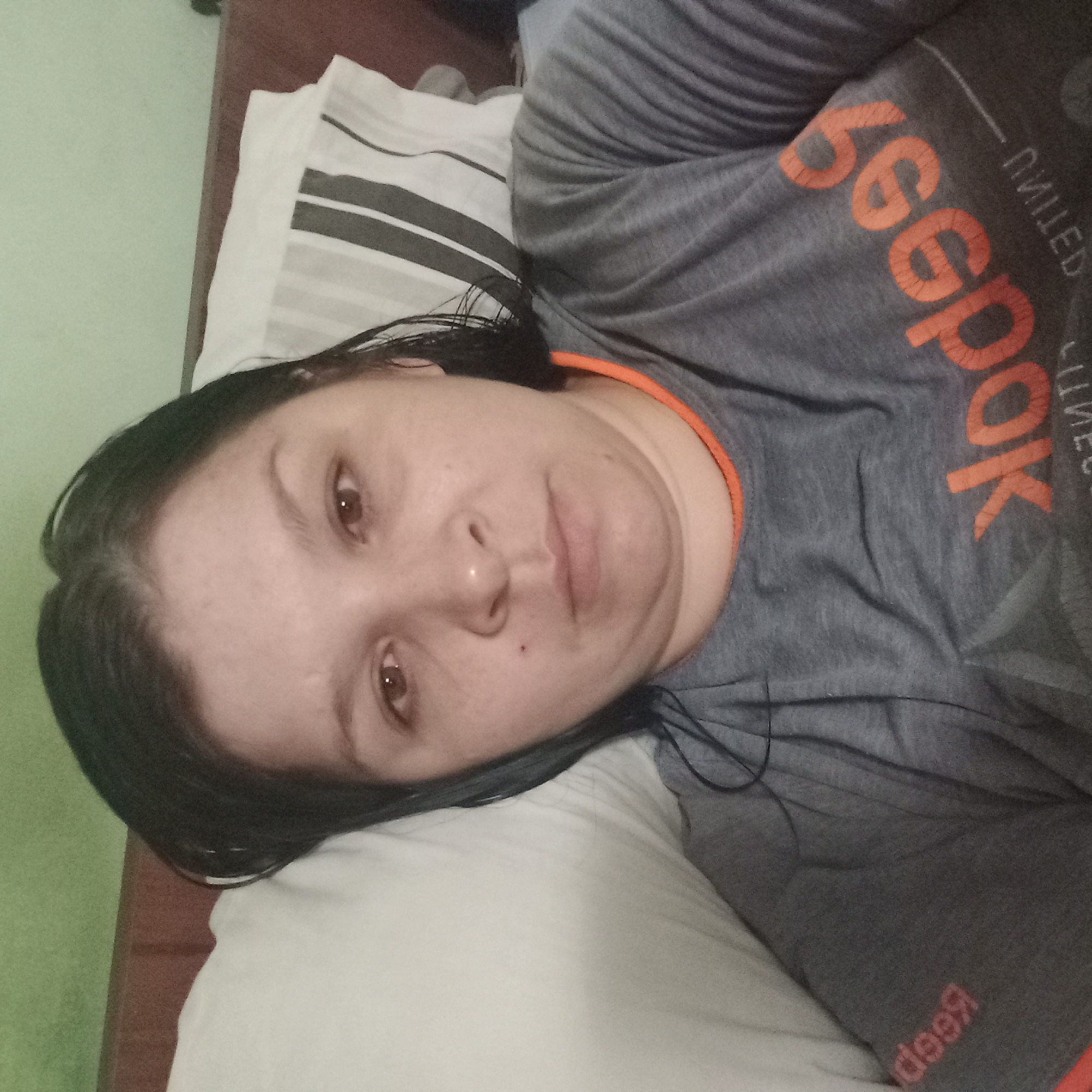 A woman with dark hair and brown eyes is lying on a bed, facing the camera. She is wearing a gray t-shirt with orange trim and an orange Reebok logo across the chest. A white pillow with dark stripes is behind her head, and a dark headboard is visible above that. The wall behind the headboard is green. The overall lighting suggests an indoor setting, possibly a bedroom, with soft, ambient light.