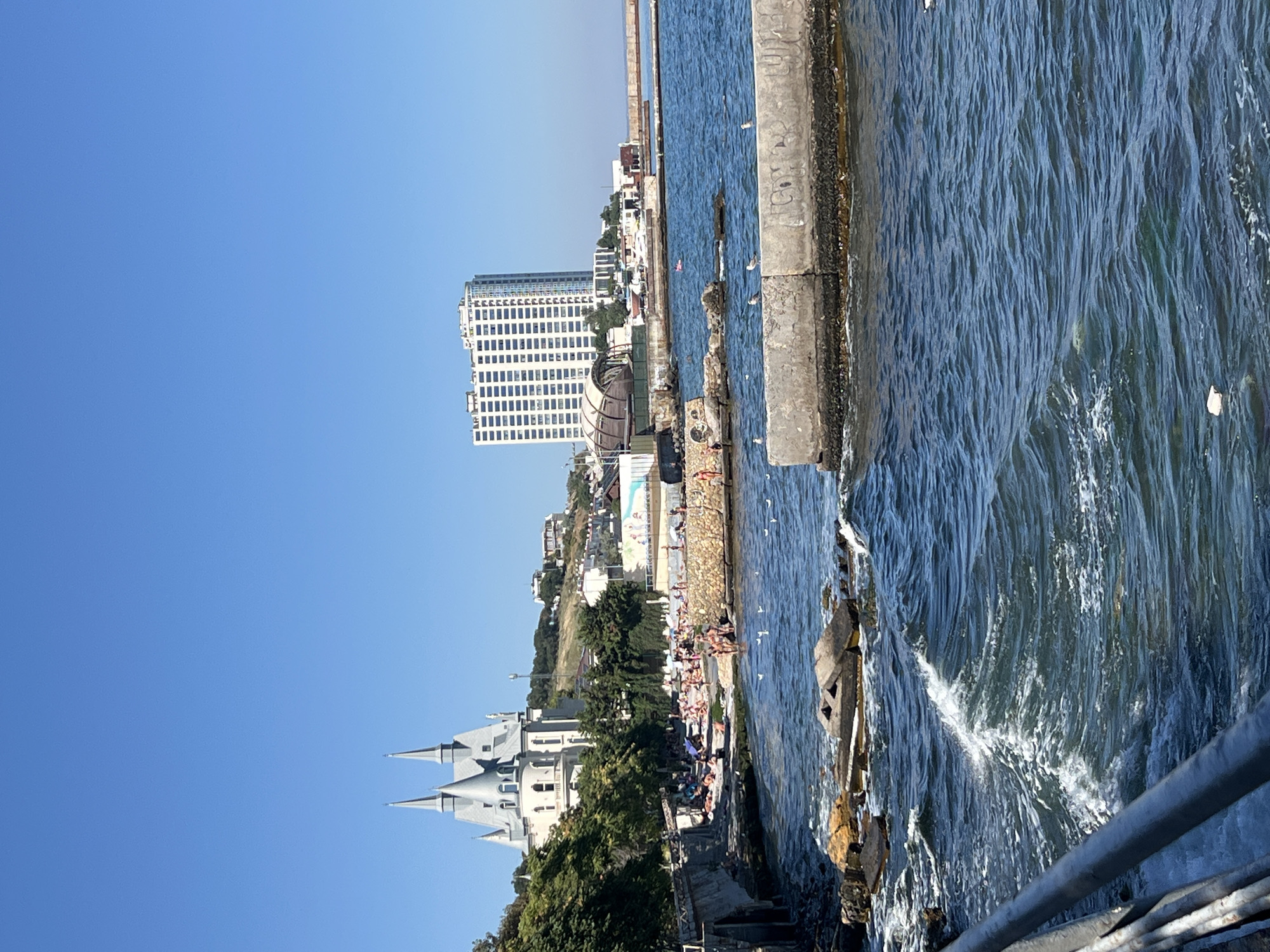 This is a wide, eye-level, horizontal shot of the Odesa coast on a sunny day. On the left, a distinctive white building with multiple pointed turrets, resembling a castle or gothic-style structure, stands on a hill. Below the building, a section of the coastline is populated by numerous sunbathers. A stone embankment separates the populated area from the turbulent, dark blue water. In the foreground, the choppy water crashes against rocks and submerged concrete structures, with seagulls dotting the surface. Further to the right, a tall, modern, multi-story white building with numerous windows dominates the skyline, situated behind a concrete pier. Another, smaller, curved structure with a white roof is visible between the castle-like building and the modern high-rise. The sky is a clear, bright blue, and the sun is high, casting strong light and shadows. The scene appears to be a popular beach area in Odesa, Ukraine, likely the coastline near the Health Path.