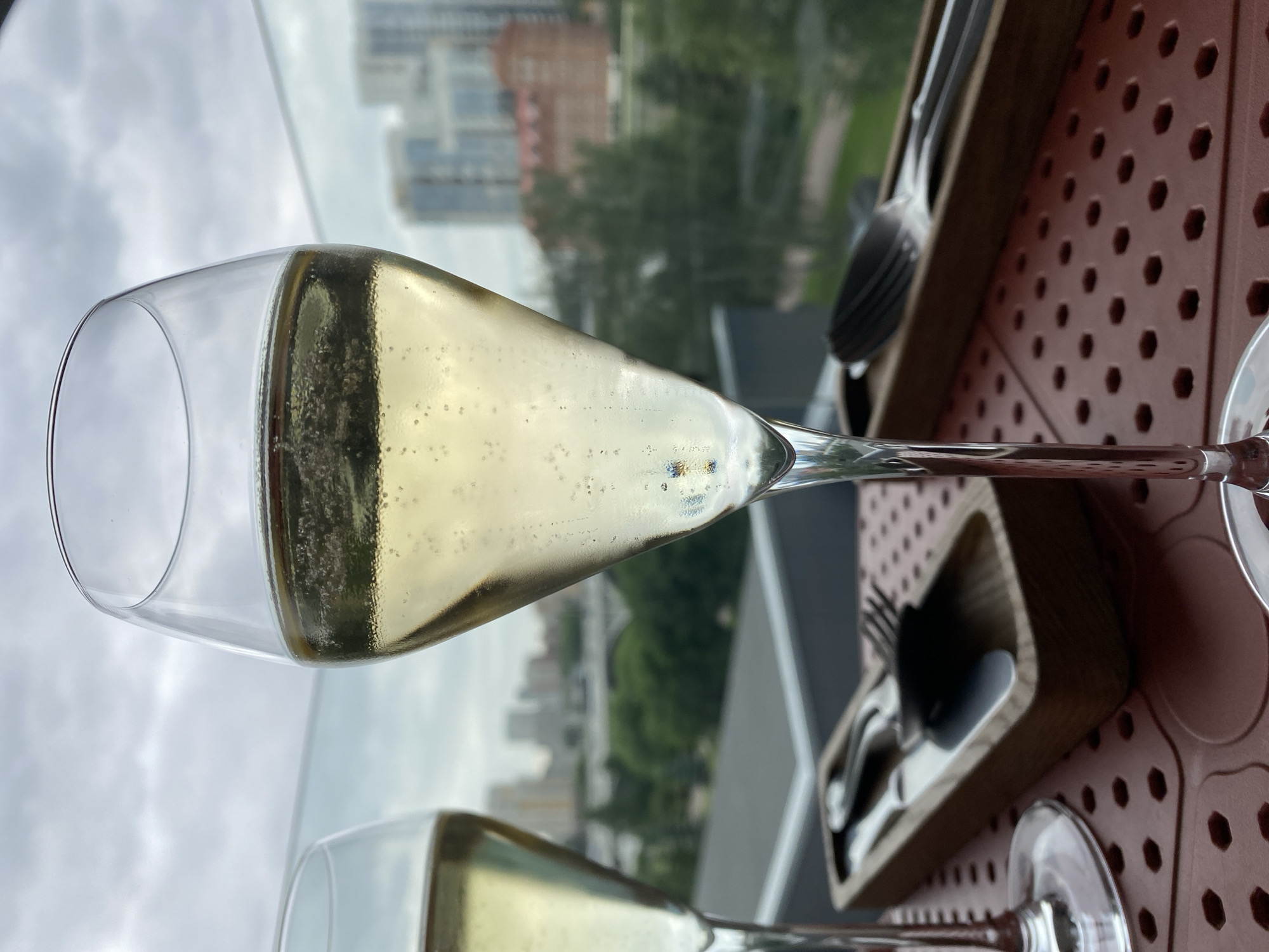 This is a close-up, eye-level, outdoor shot of two champagne flutes filled with a light-colored beverage, likely champagne or sparkling wine, with visible bubbles. The flutes are positioned on a table with a patterned surface. In the foreground, there are two wooden trays containing silverware, one holding a fork and knife, the other a spoon and knife.
In the background, a blurred cityscape is visible, suggesting an urban setting. The buildings appear to be modern, with some older architecture also present. Green trees and foliage are scattered throughout the urban landscape. The sky is overcast with clouds.
Based on the provided context, this scene is set in Yekaterinburg, Russia. The view suggests the location is likely a restaurant, bar, or rooftop terrace offering a view of the city. There are no people visible in the image. The lighting suggests it is daytime, but the cloudy sky gives a soft, diffused light.
