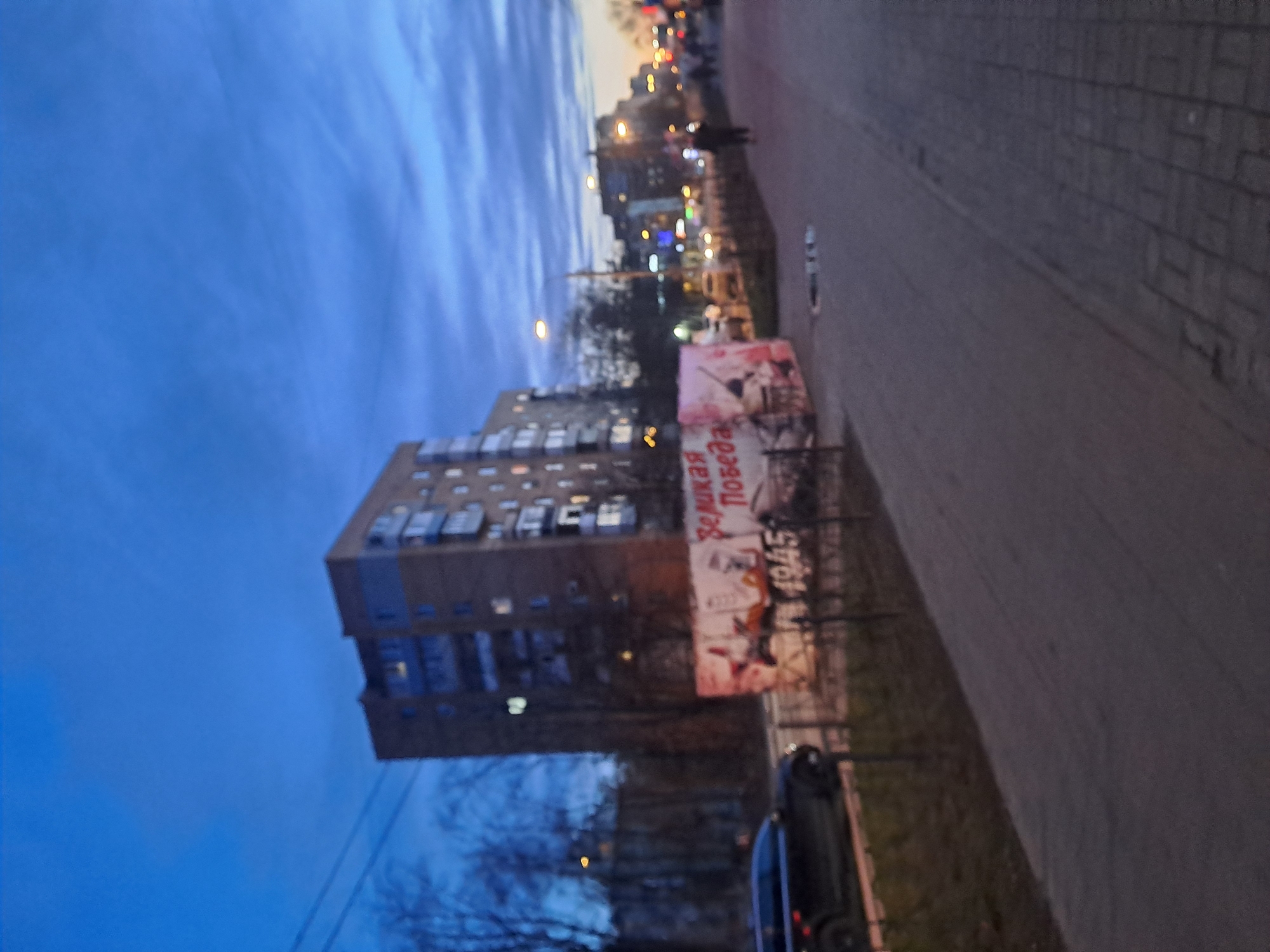 The image depicts a street scene at dusk in Elektrostal, Russia. A multi-story apartment building is visible on the left side of the frame. Adjacent to the building is a roadside advertisement displaying a "Great Victory 1945" poster. A dark-colored car is parked nearby, and a sidewalk runs along the right side of the image. Streetlights are visible in the distance, along with silhouettes of people walking. The sky is a blend of blues.