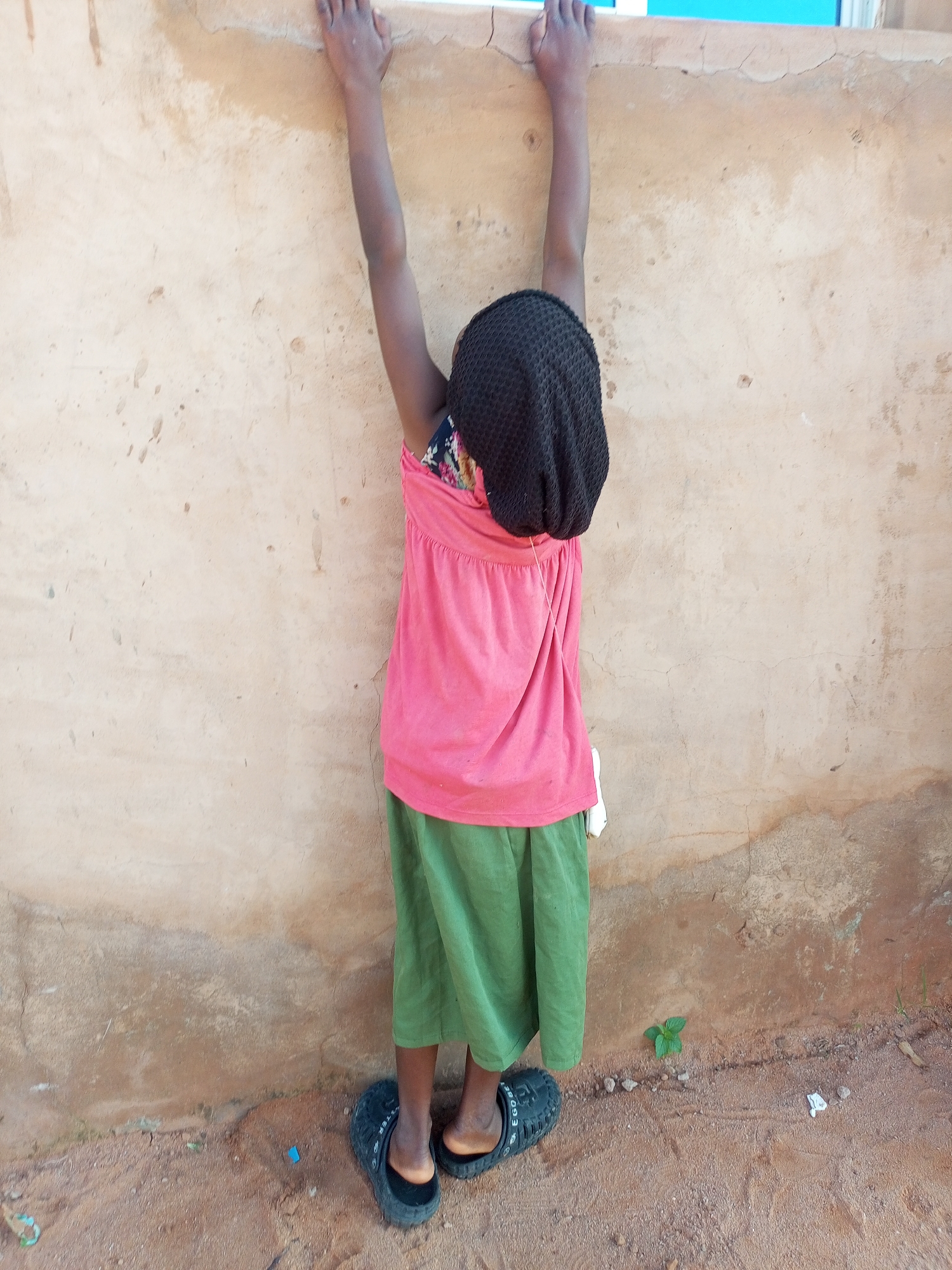 A young girl stands with her arms raised and clasped to a textured, light brown wall. She is wearing a black, close-fitting hat, a pink sleeveless top with a patterned insert around the neckline, and a long, green skirt. Her feet are clad in black, slip-on sandals. The ground beneath her is sandy and dusty, with a few small pebbles and a tiny green plant growing near the wall. The wall has some cracks and discoloration, suggesting it is an outdoor surface, possibly part of a building in a rural or semi-urban area of Tempure, Nigeria.