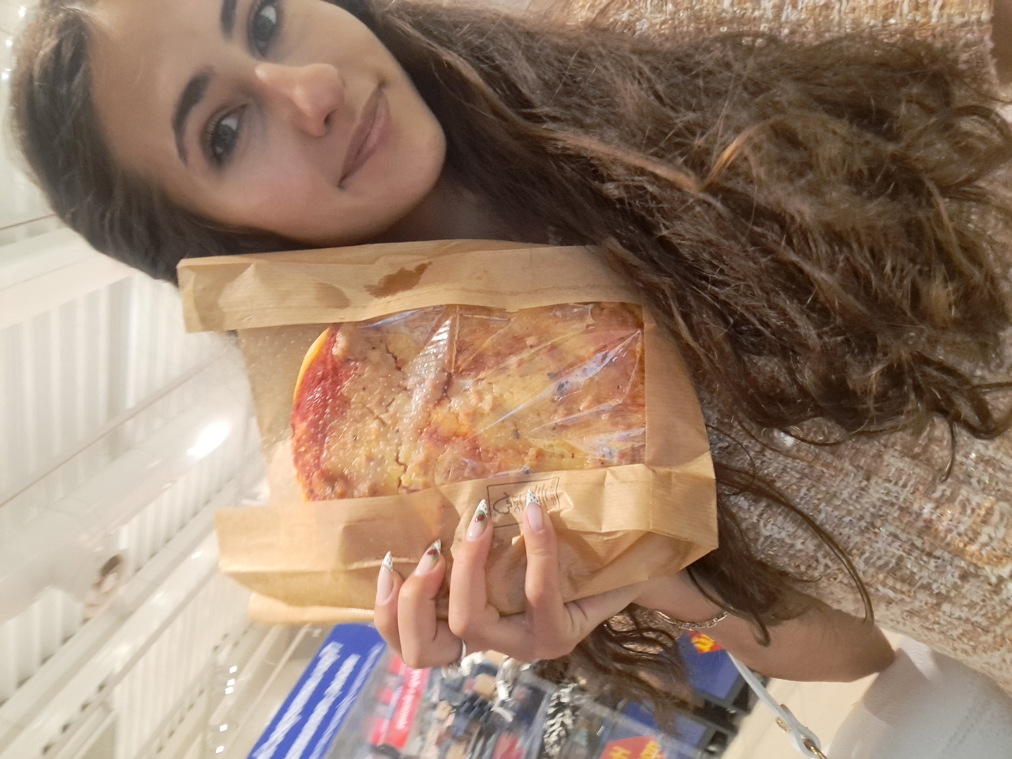 A woman with long brown hair and patterned fingernails holds a rectangular pizza in a brown paper bag. The pizza is encased in clear plastic. The background appears to be a store shelf, suggesting the image was taken in a shop. No information suggests the location to be Sofia, Bulgaria.
