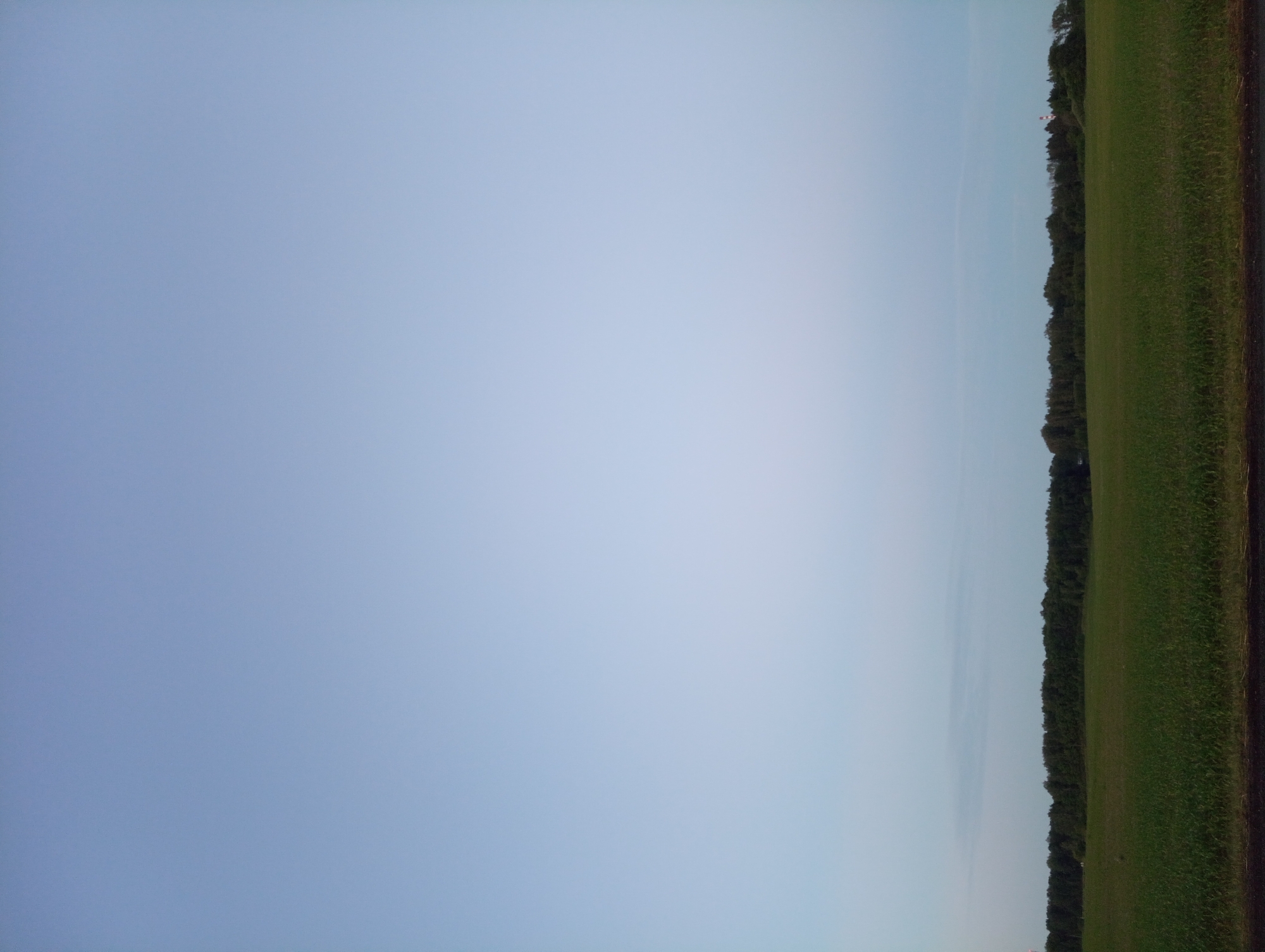 The image shows a wide, flat expanse of farmland with a dark horizon line of trees. The sky is a pale, hazy blue, suggesting either early morning or late evening. In the distance, on the right side of the image, a red and white striped industrial smokestack is visible above the trees. The foreground consists of a green field, likely crops, and a dark, indistinct border along the bottom of the frame. There are no people or animals visible. The scene appears to be located in the rural area of Палыкавіцкі сельскі Савет, Belarus.