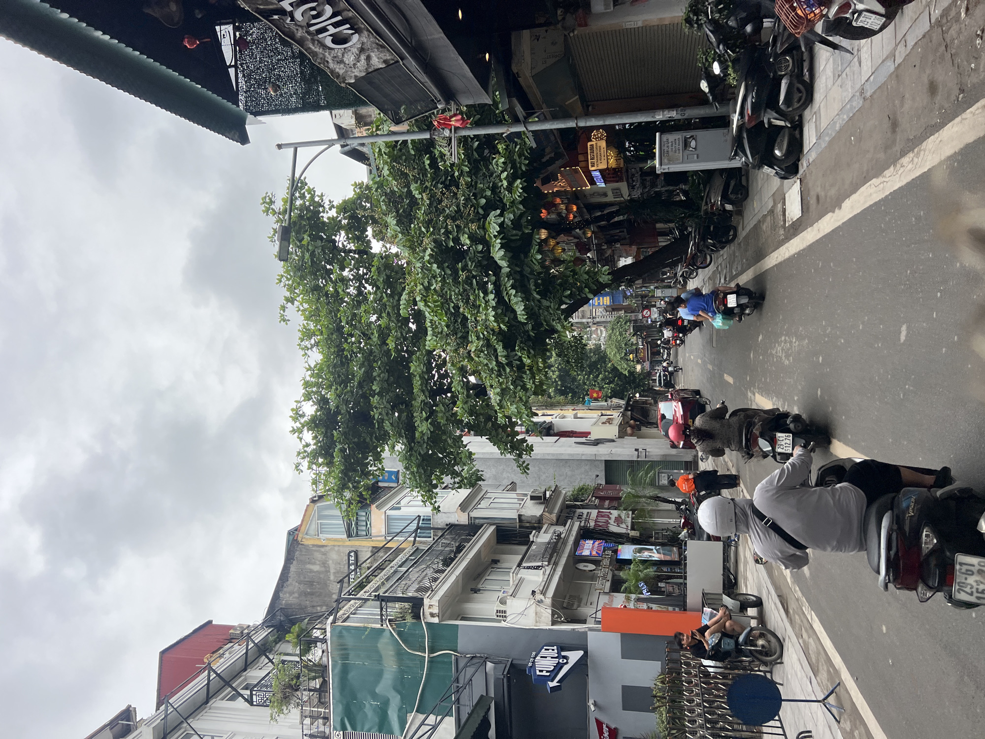 This is a medium-angle, eye-level shot of a street in Hanoi, Vietnam, on a cloudy day. The scene captures the bustling activity of a city street, dominated by motorbikes and narrow buildings.
In the foreground, three motorbikes are visible on the asphalt road. The closest motorbike has a rider wearing a light gray long-sleeved shirt, a white helmet, and dark pants, with a black bag slung across their chest. The motorbike's rear license plate reads "29-G1 152.29". Behind this motorbike, another rider on a dark motorbike is partially visible, with a license plate showing "29-C1 512.76". Further down the road, a third motorbike carries a person in a blue shirt and a helmet.
On the right side of the street, a building with a dark green, corrugated metal roof overhangs the sidewalk. Signs and lanterns are visible on the building's facade, including one that appears to say "CHỢ". Parked motorbikes line the sidewalk.
On the left side of the street, a series of multi-story buildings are lined up. These buildings vary in architectural style and color, with balconies, windows, and awnings. One building has a prominent blue sign with white lettering that reads "FUNFUEL COCO CO". A person is sitting on a motorbike parked on the sidewalk next to a white cart. Another motorbike with a rider is parked slightly further back.
A large tree with dense green foliage stands in the middle of the street, extending upwards towards the sky, partially obscuring the buildings behind it. The sky is overcast, with thick, gray clouds. The overall atmosphere is that of a typical busy urban environment in Southeast Asia.