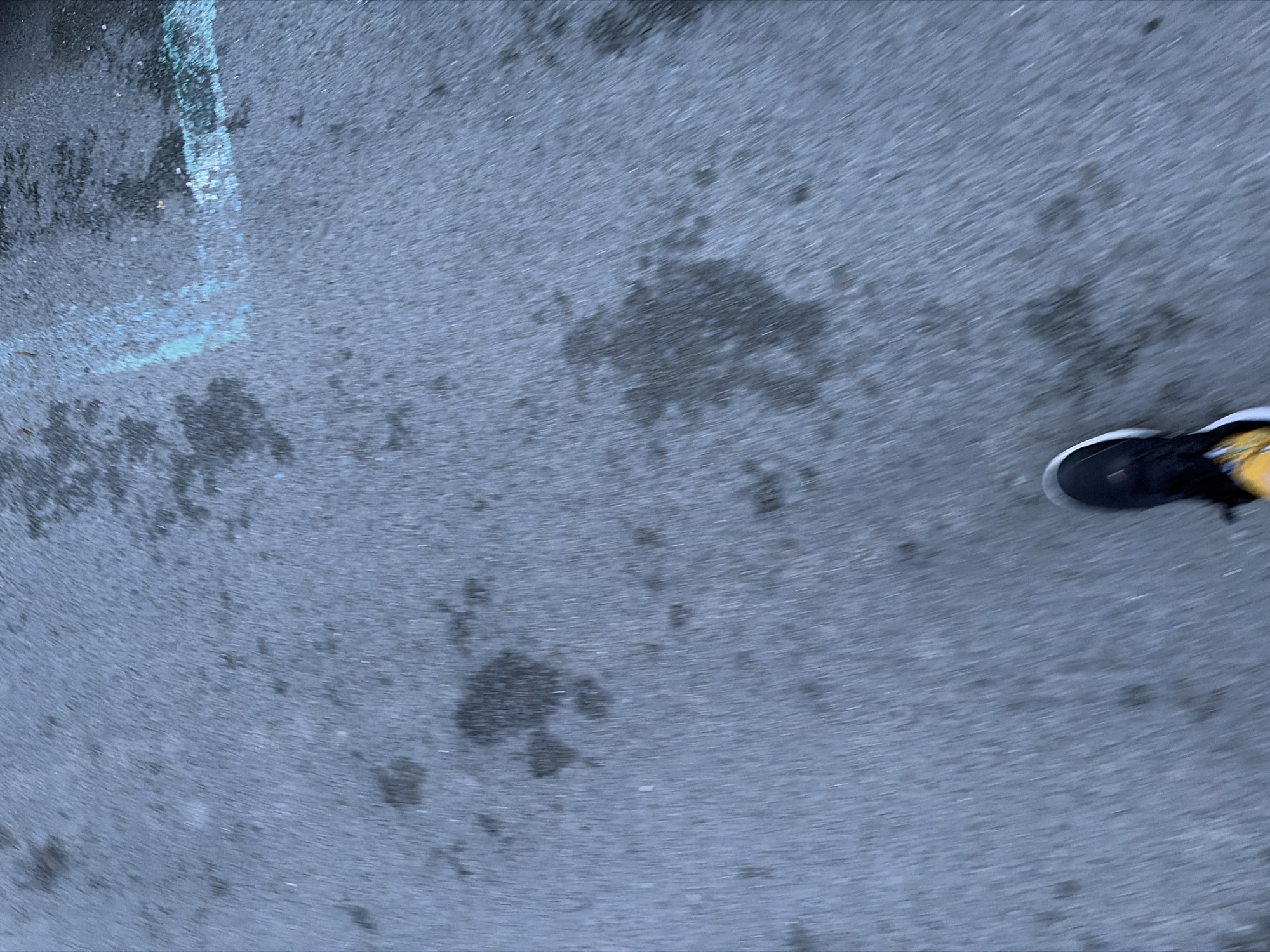 The image presents an overhead, motion-blurred view of an asphalt surface with a partially visible foot in the lower right corner. The foot is wearing a black sneaker with yellow accents and a white sole. The ground is dark grey asphalt, marked with irregular dark patches that suggest wetness or stains. In the upper left portion of the image, there are faint, light blue chalk markings, appearing as a worn line or abstract shape on the pavement. The strong horizontal motion blur across the entire frame indicates movement, likely from the person taking the photograph while walking. The setting is an outdoor ground surface, possibly a street, sidewalk, or parking lot in Burgas, Bulgaria. There is no visible text or specific location cues beyond the ground type and chalk.